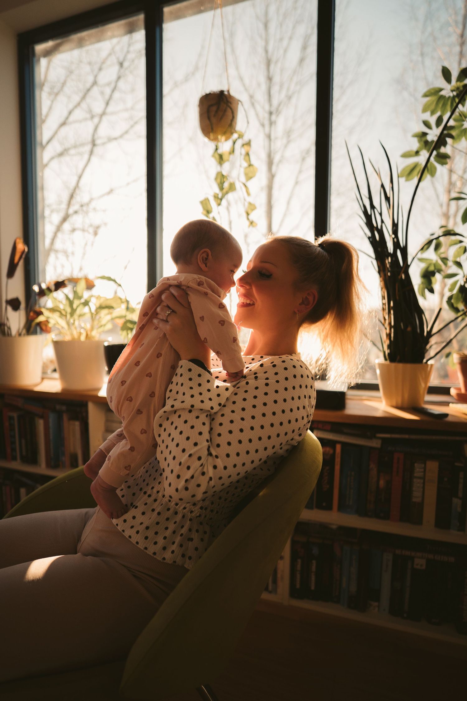 Mother holding newborn baby near sunlit window with plants and bookshelves in cozy home interior.