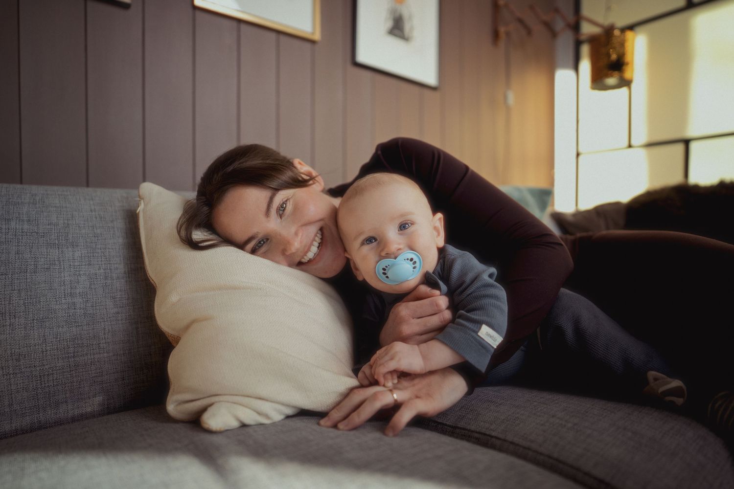 A mother tenderly cuddles her baby with a blue pacifier on a cozy gray sofa at home.