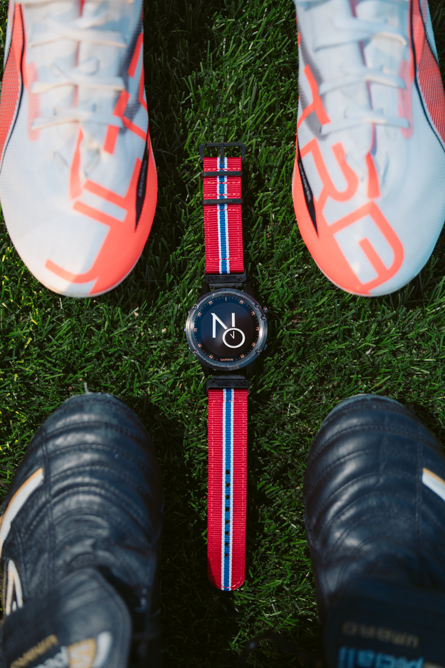 A stylish watch with red, white, and blue NATO strap placed on grass between two pairs of soccer cleats.