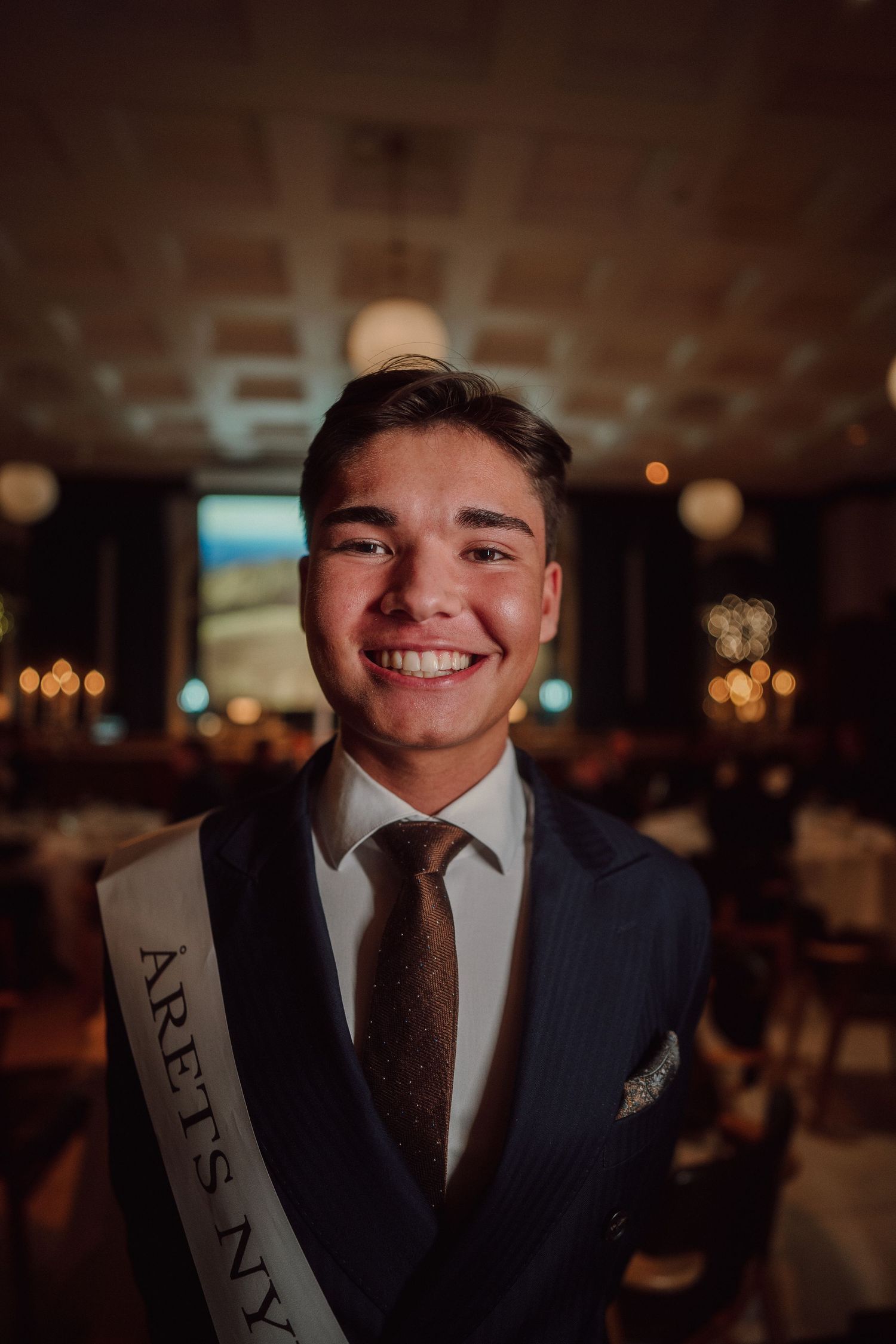 Person in formal attire wearing celebration sash smiling at evening event.
