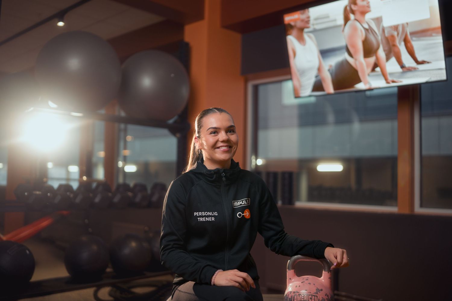 Smiling woman in a gym wearing a black jacket, sitting near kettlebells with a fitness screen in the background.
