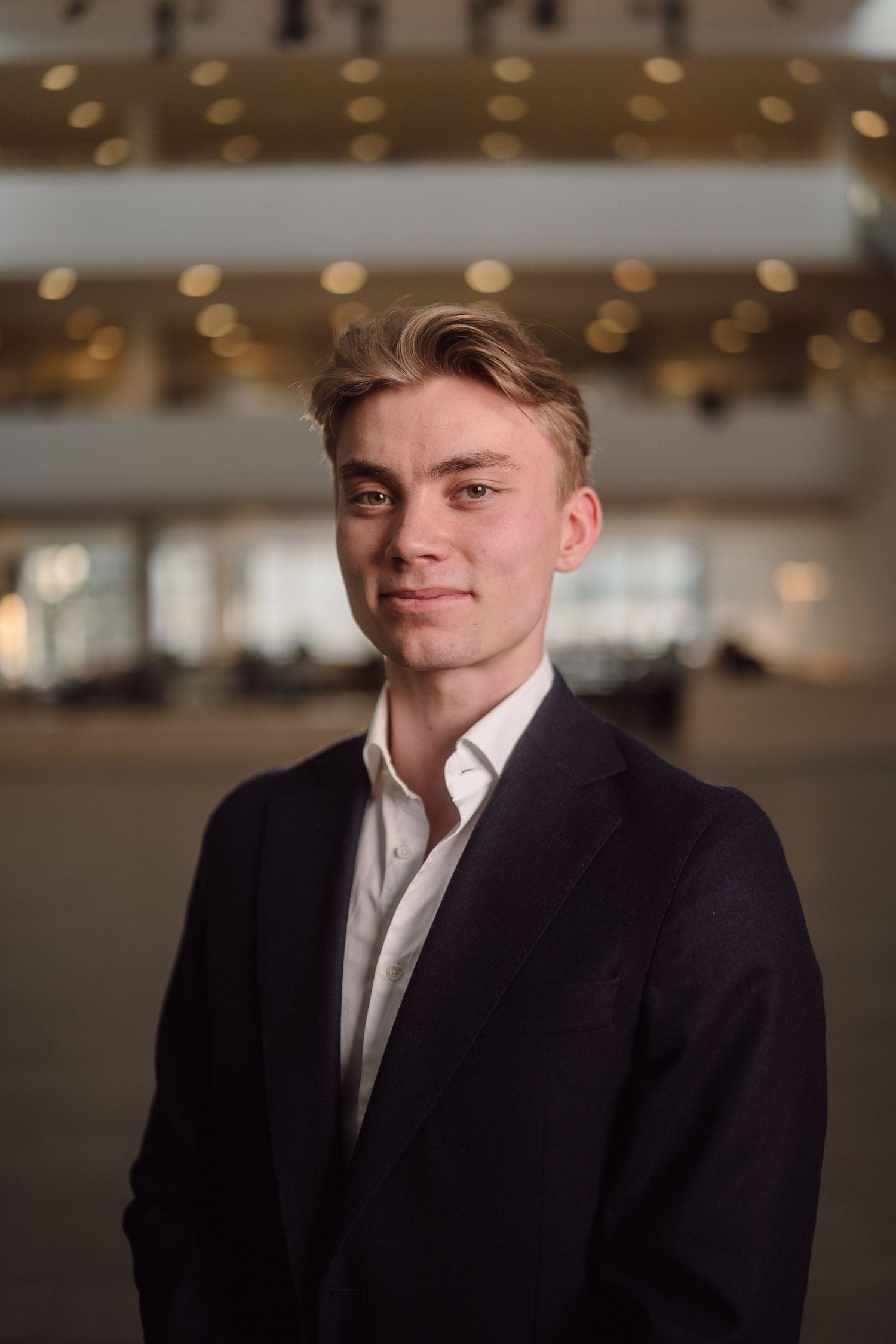 A professional headshot of someone in business attire against an indoor background with ambient lighting.
