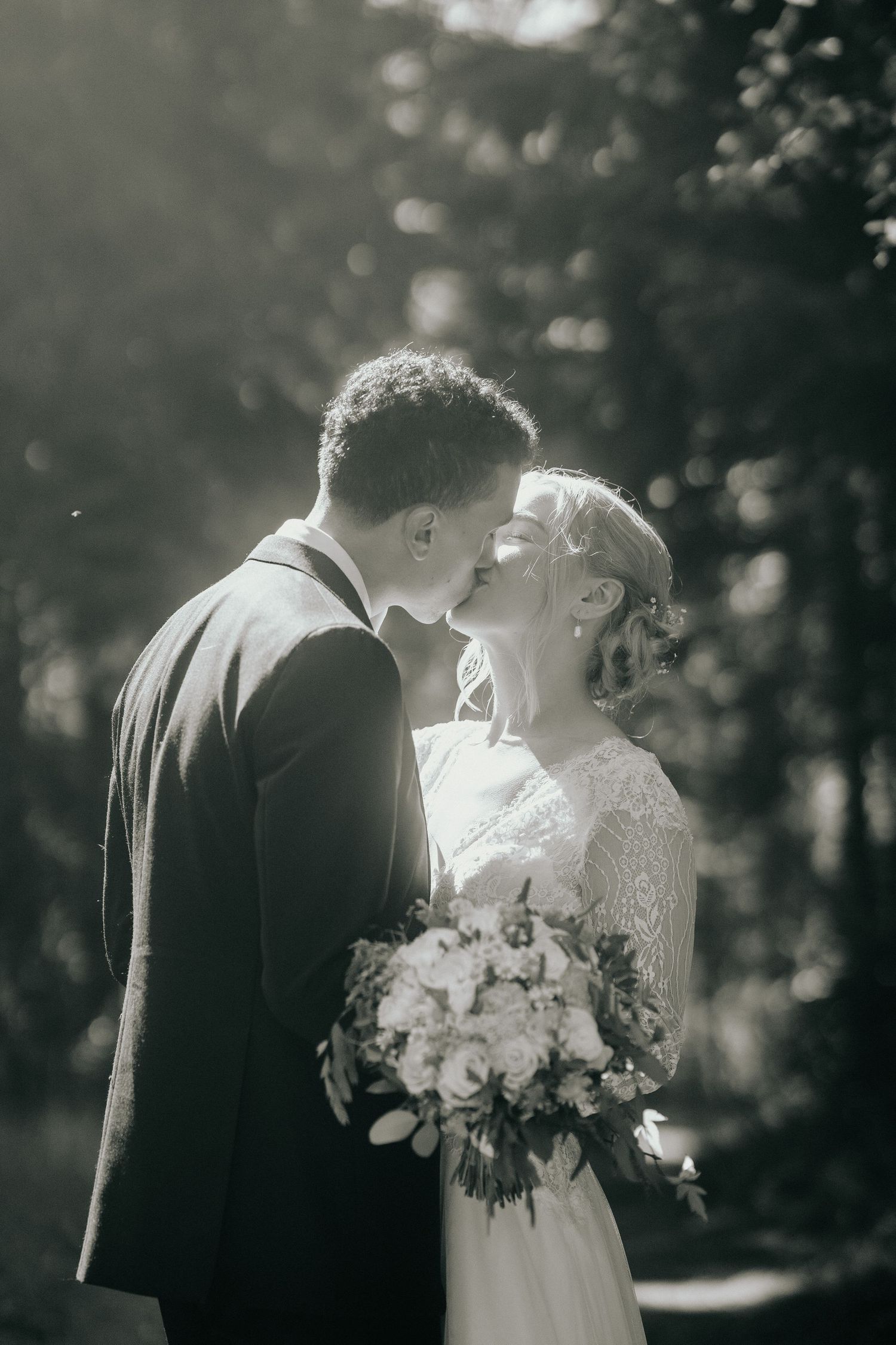 Black and white romantic wedding photo of a couple kissing while holding a floral bouquet in a forest setting.