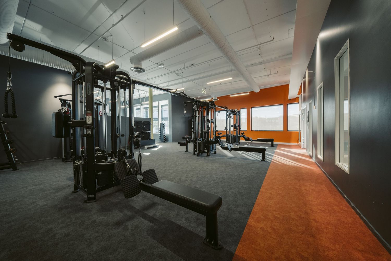 Modern gym interior with black cable machines, weight equipment, and orange accent walls on gray carpet flooring.