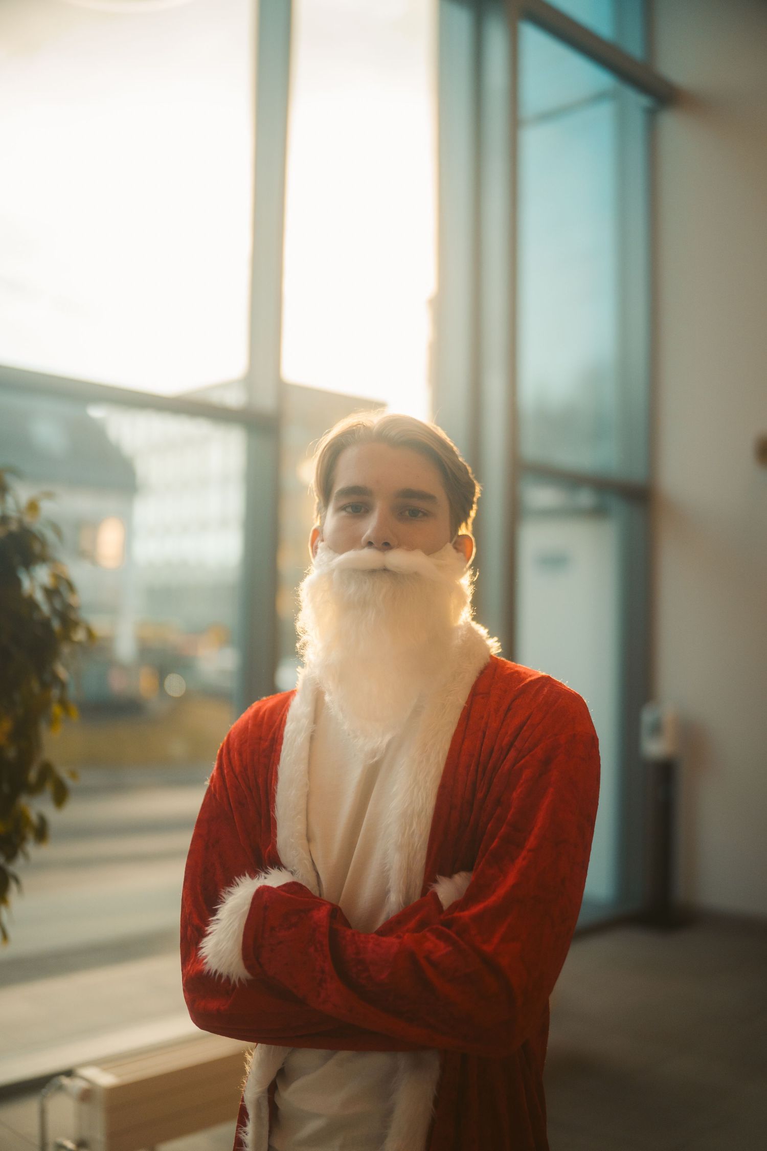 Person wearing Santa Claus costume with white beard and red cardigan standing by window in warm lighting.