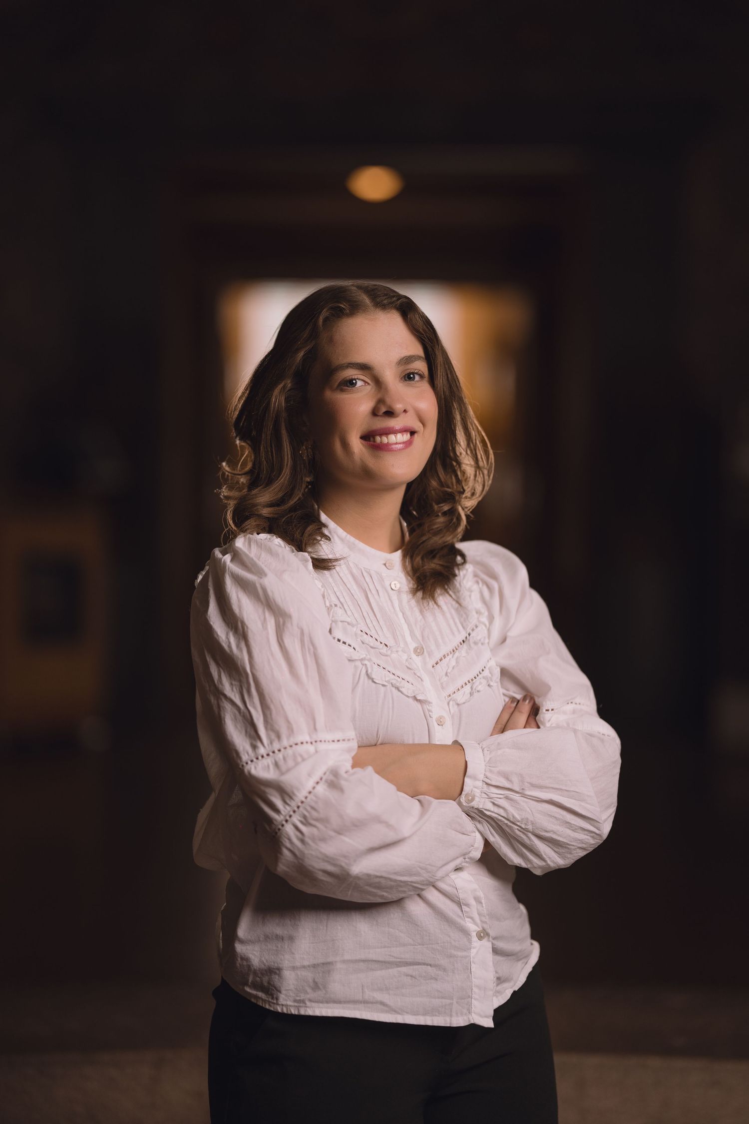 A person in a white button-down shirt crosses their arms while posing in a dramatic indoor setting.