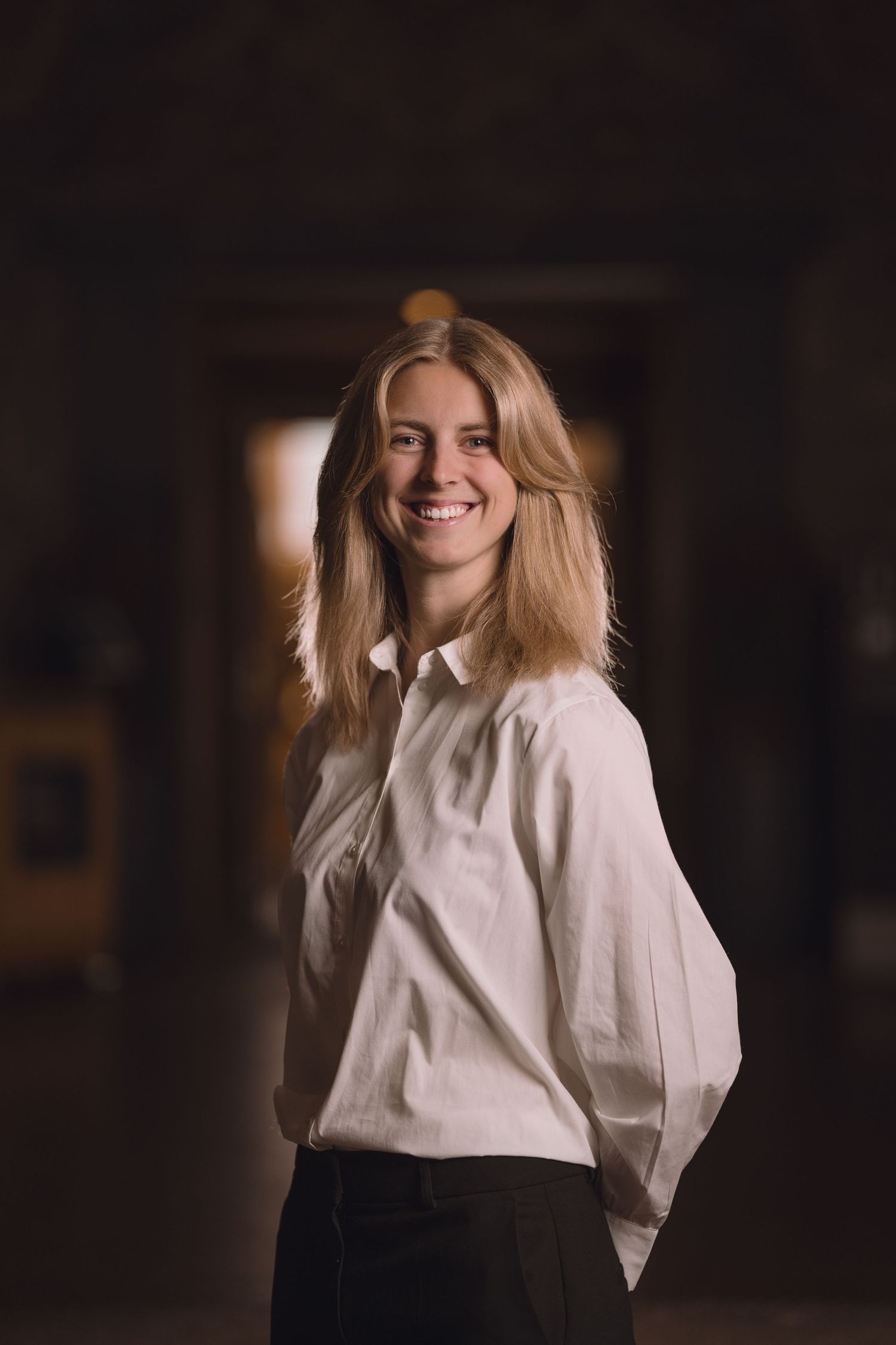 A portrait of someone in casual white attire with blonde hair against a moody dark background.