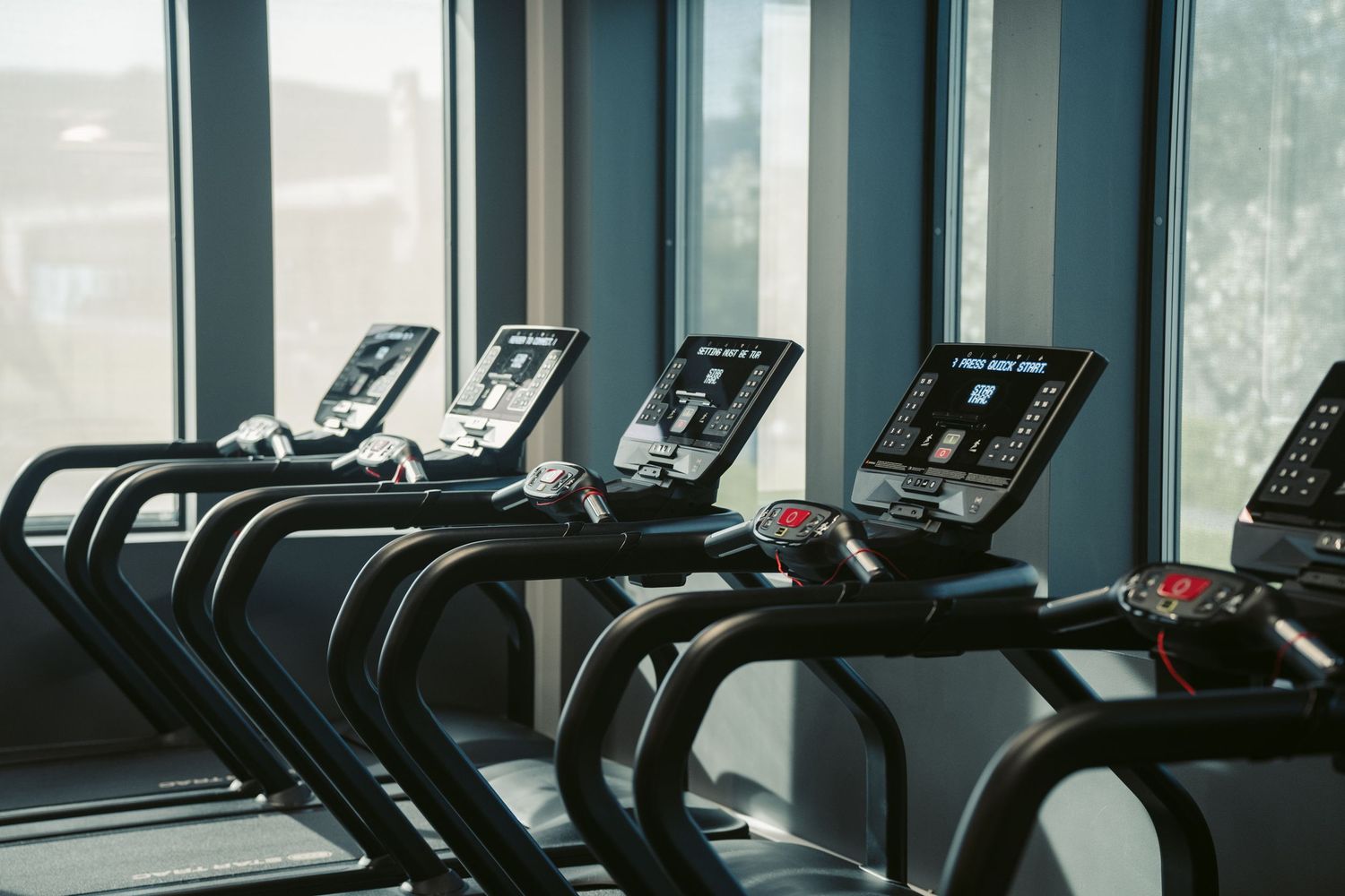 Row of modern treadmills lined up by large windows in a bright, well-equipped gym fitness center.