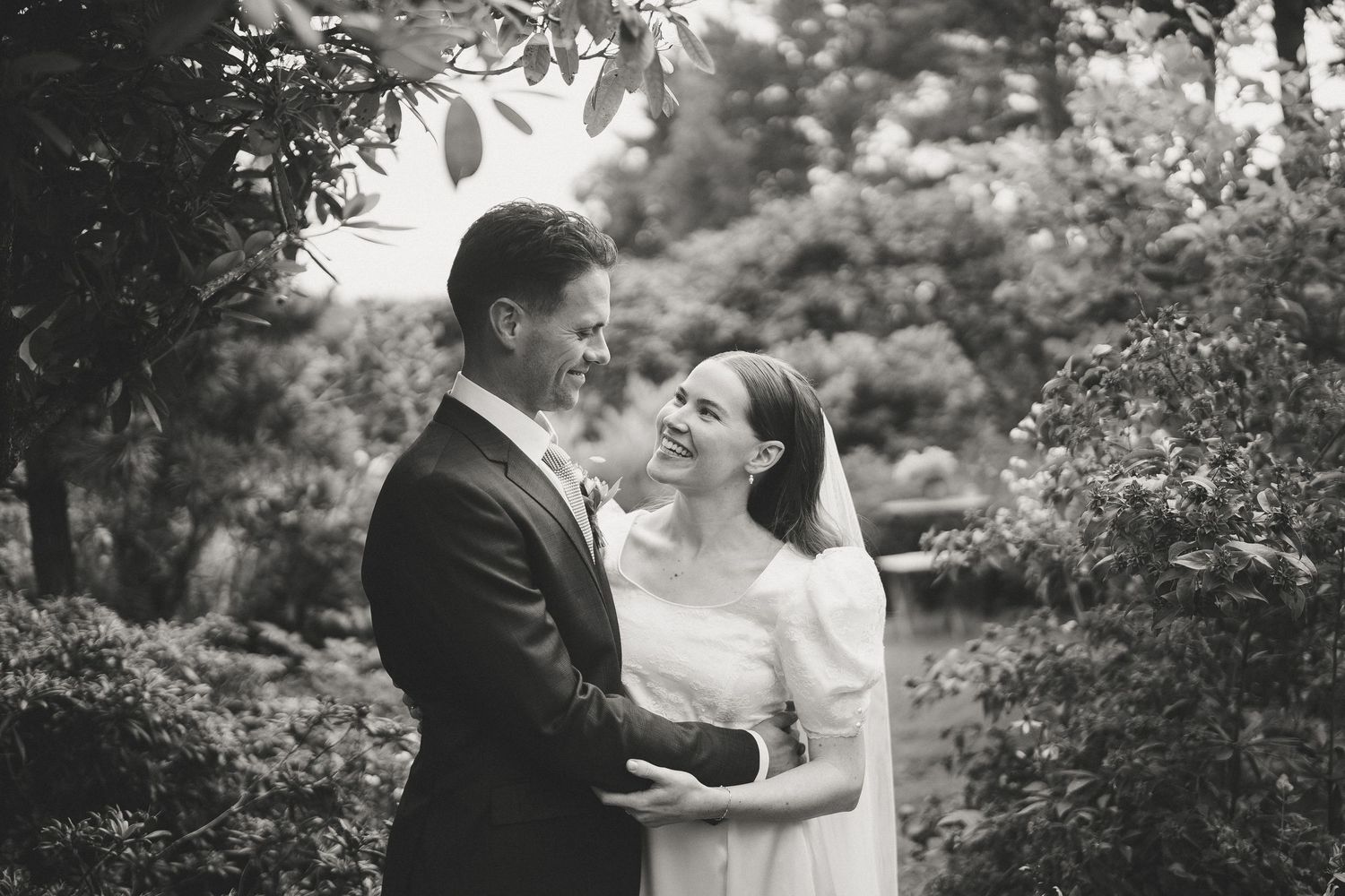 Black and white wedding photo of couple embracing in a lush garden setting.