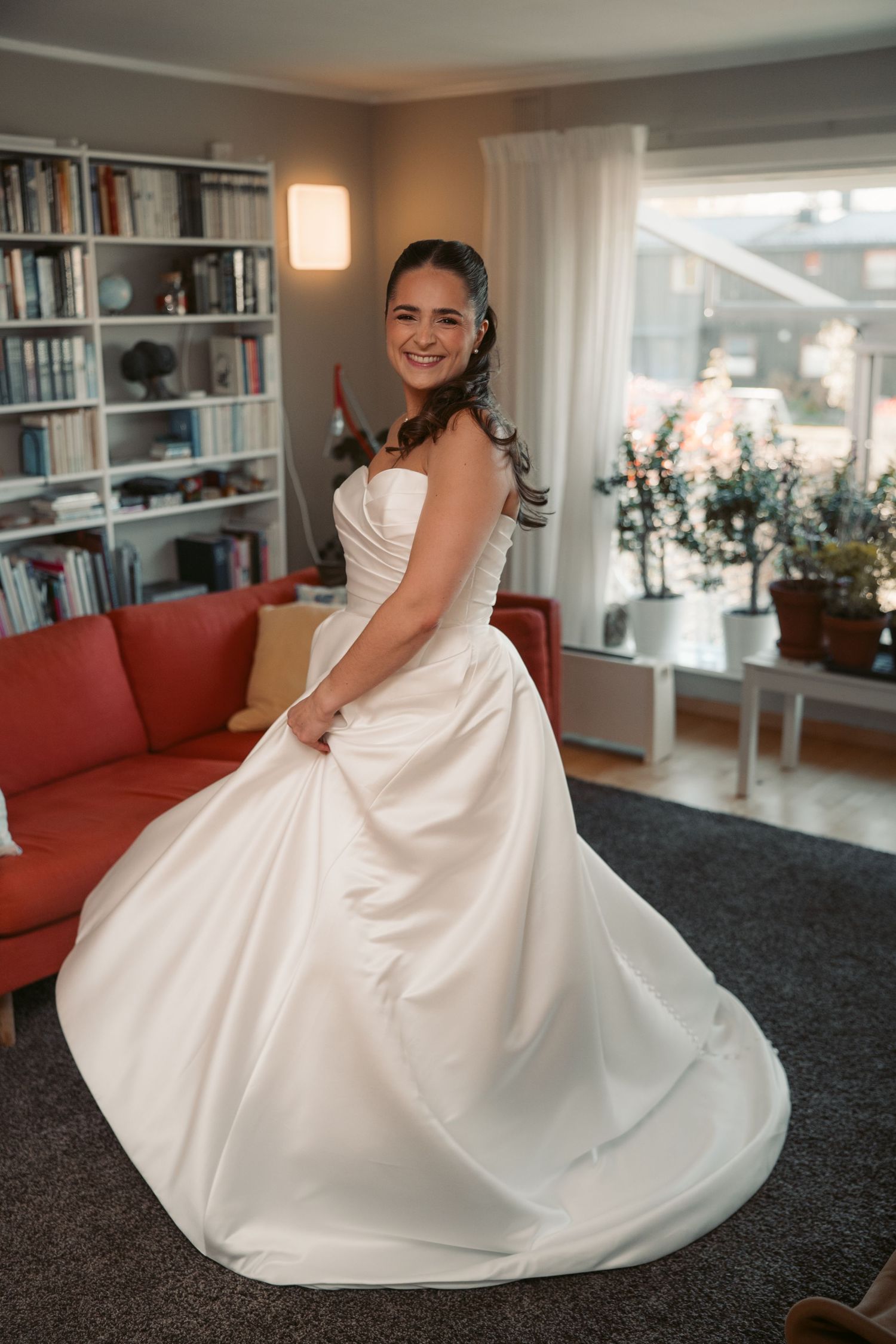 Elegant white wedding dress displayed in a modern living room with red couch and built-in bookshelves.