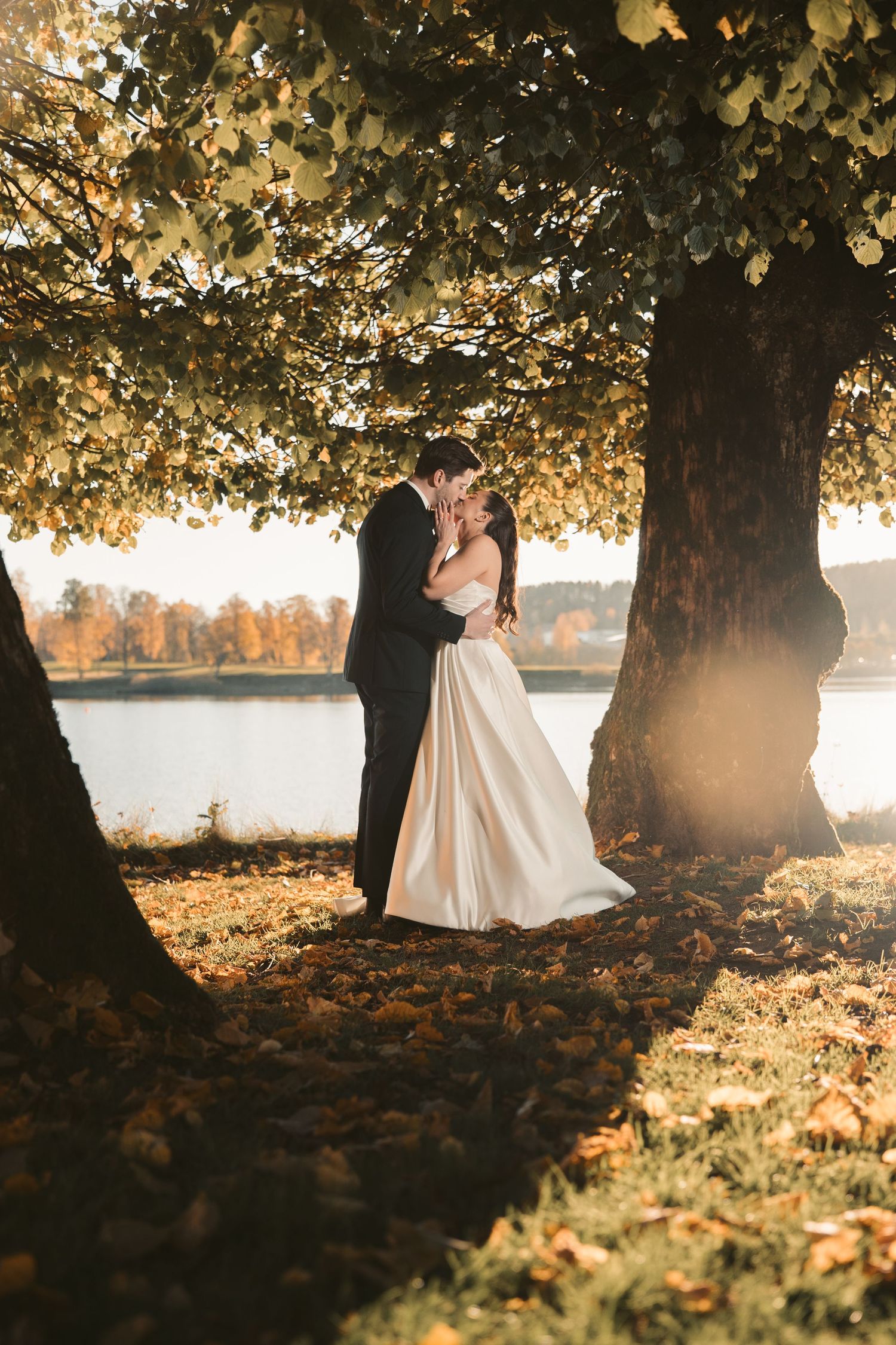 Silhouetted wedding couple under autumn trees by a lake during golden hour.