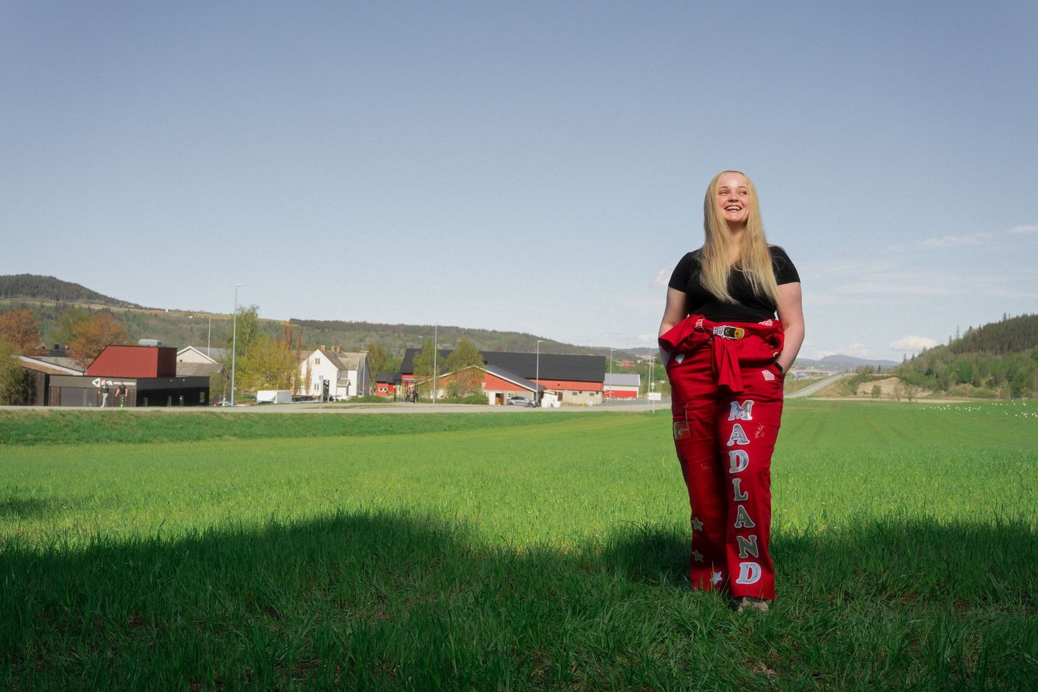 Blonde woman in red Maryland pants stands smiling in green field with red farm buildings and hills in background.