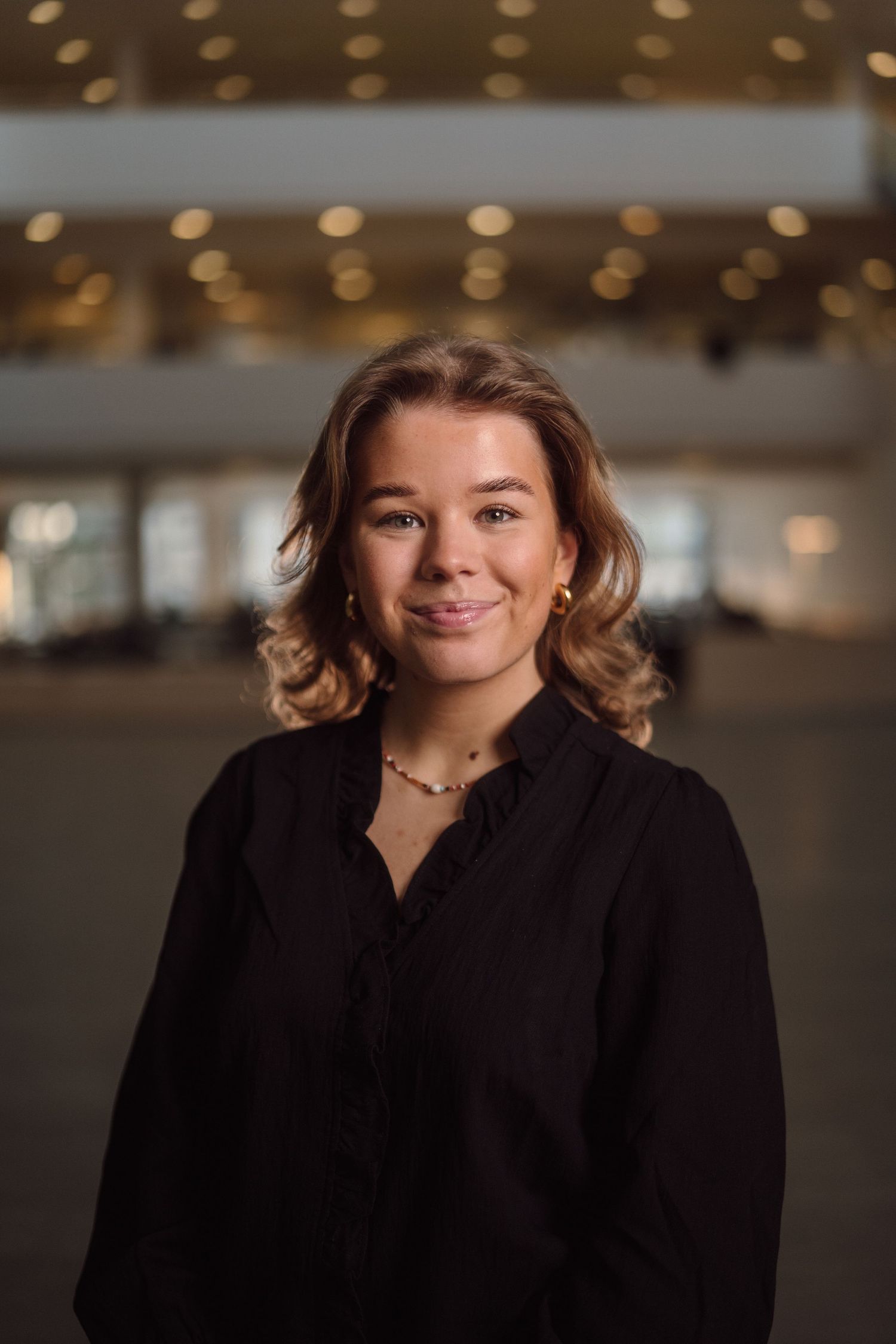 A professional portrait of someone wearing a black blazer against a warm-lit interior background.