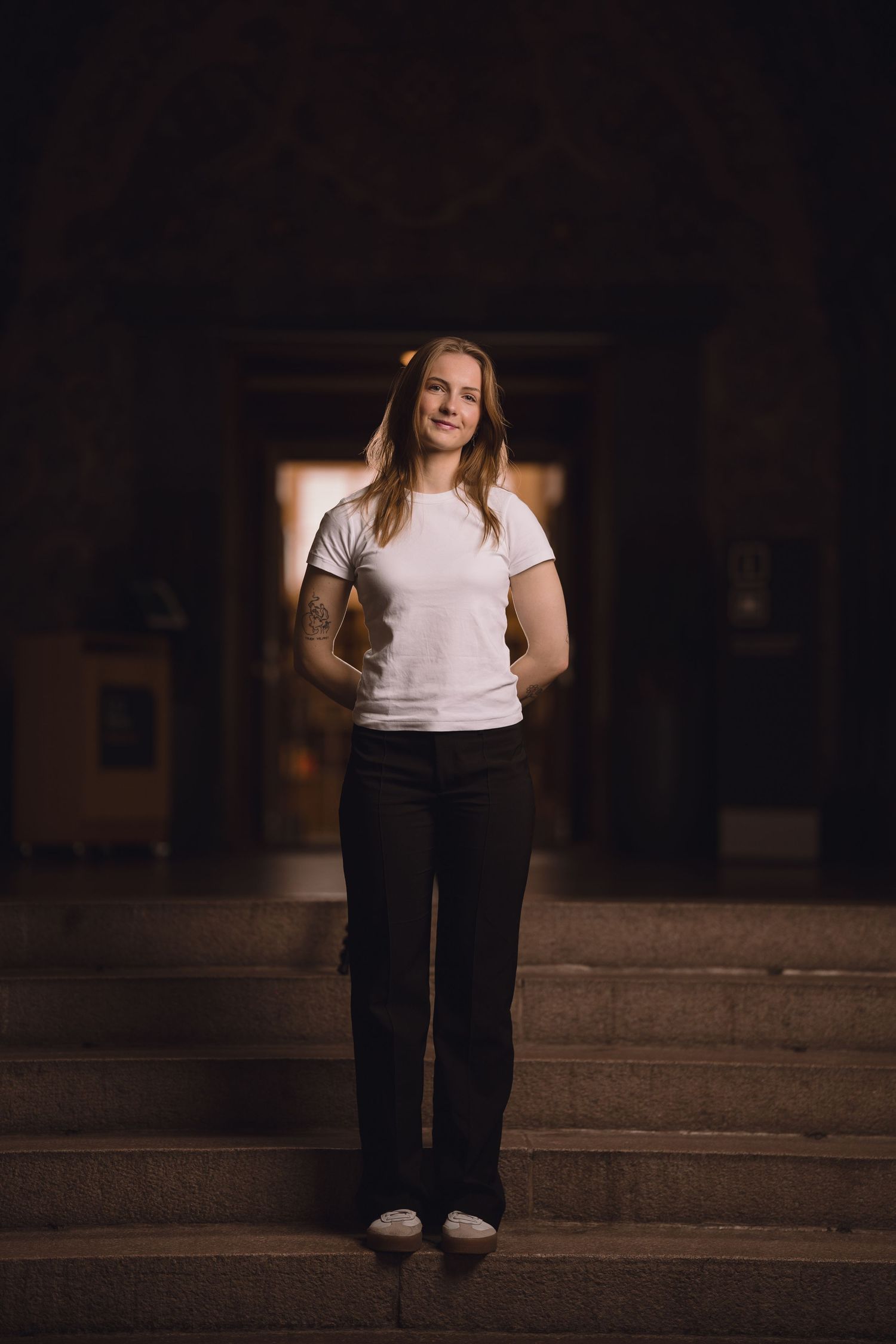 A figure wearing a white top and black pants stands in a dimly lit space with dramatic lighting.
