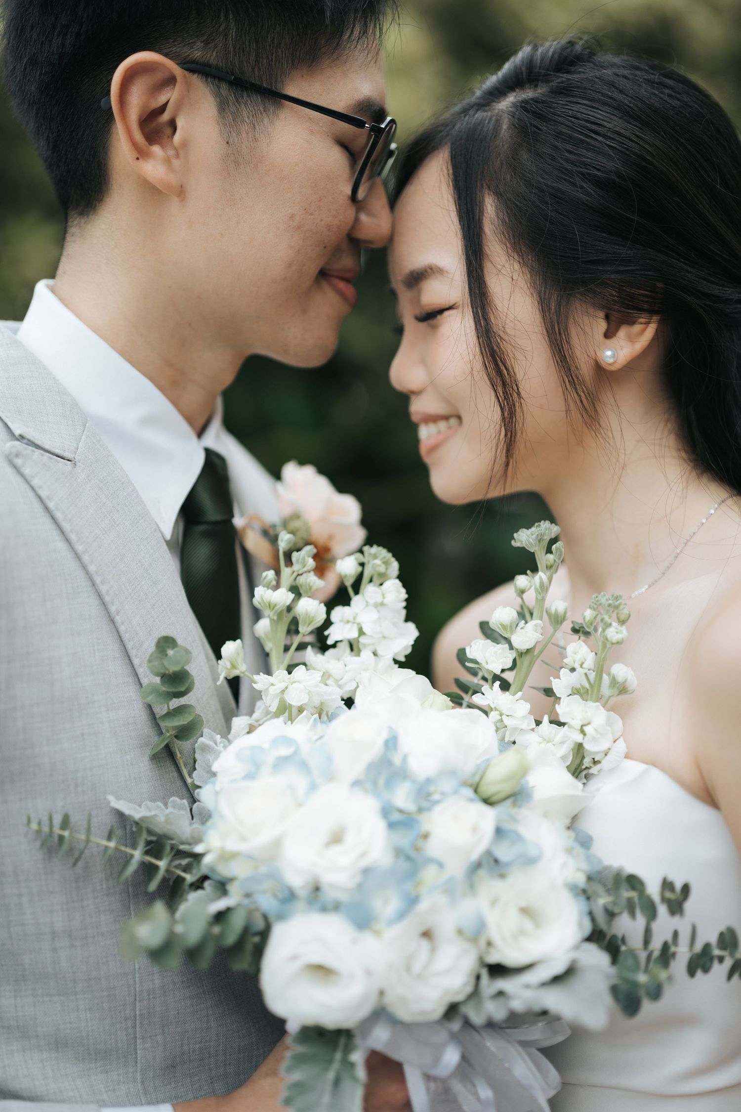 A romantic wedding couple in grey and white share intimate moments with a beautiful white floral bouquet in an outdoor setting.