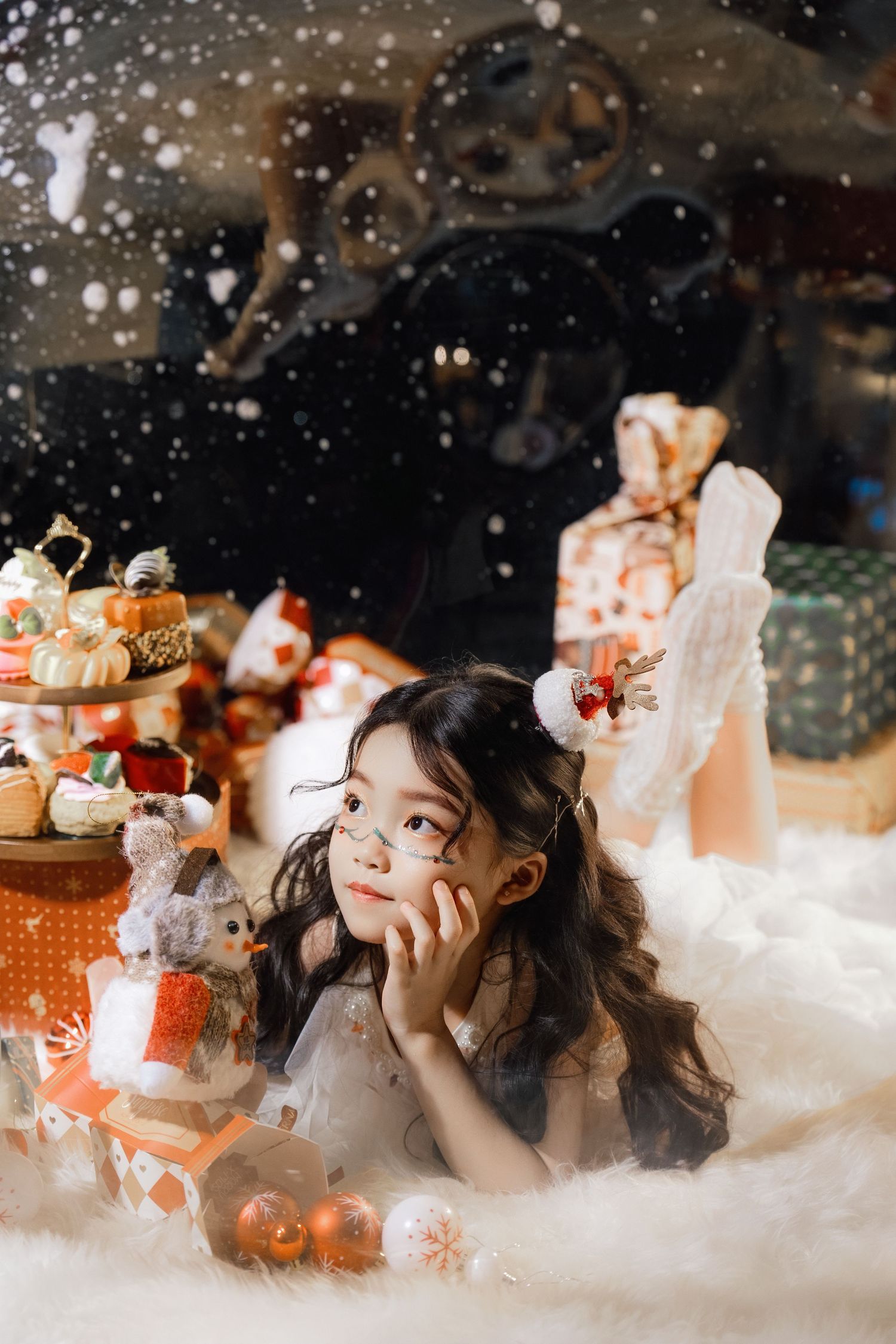 Young Asian girl lying in fake snow surrounded by Christmas gifts, toys, and decorations in a festive holiday setting.