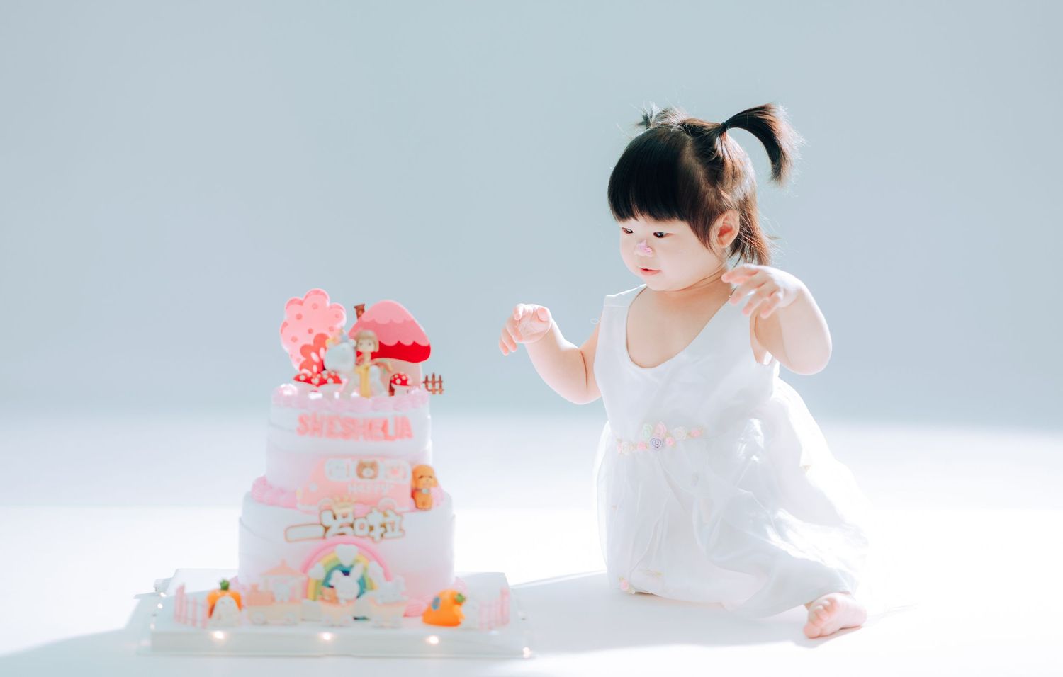 A toddler girl in a white dress sits beside a pink tiered birthday cake on a white background.