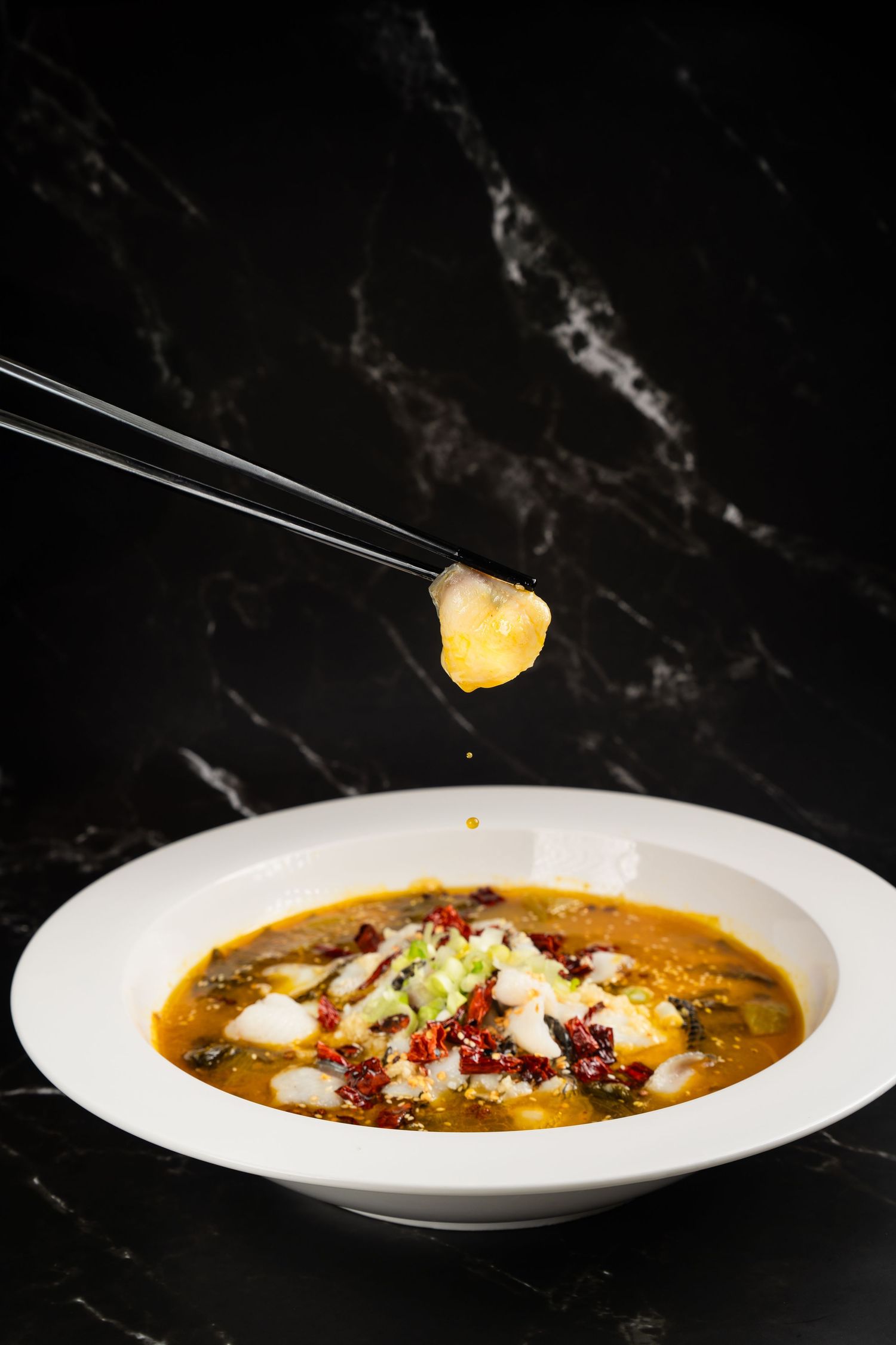 Chopsticks lifting a piece of fish from a white bowl of spicy Chinese sour soup with red peppers on black marble.