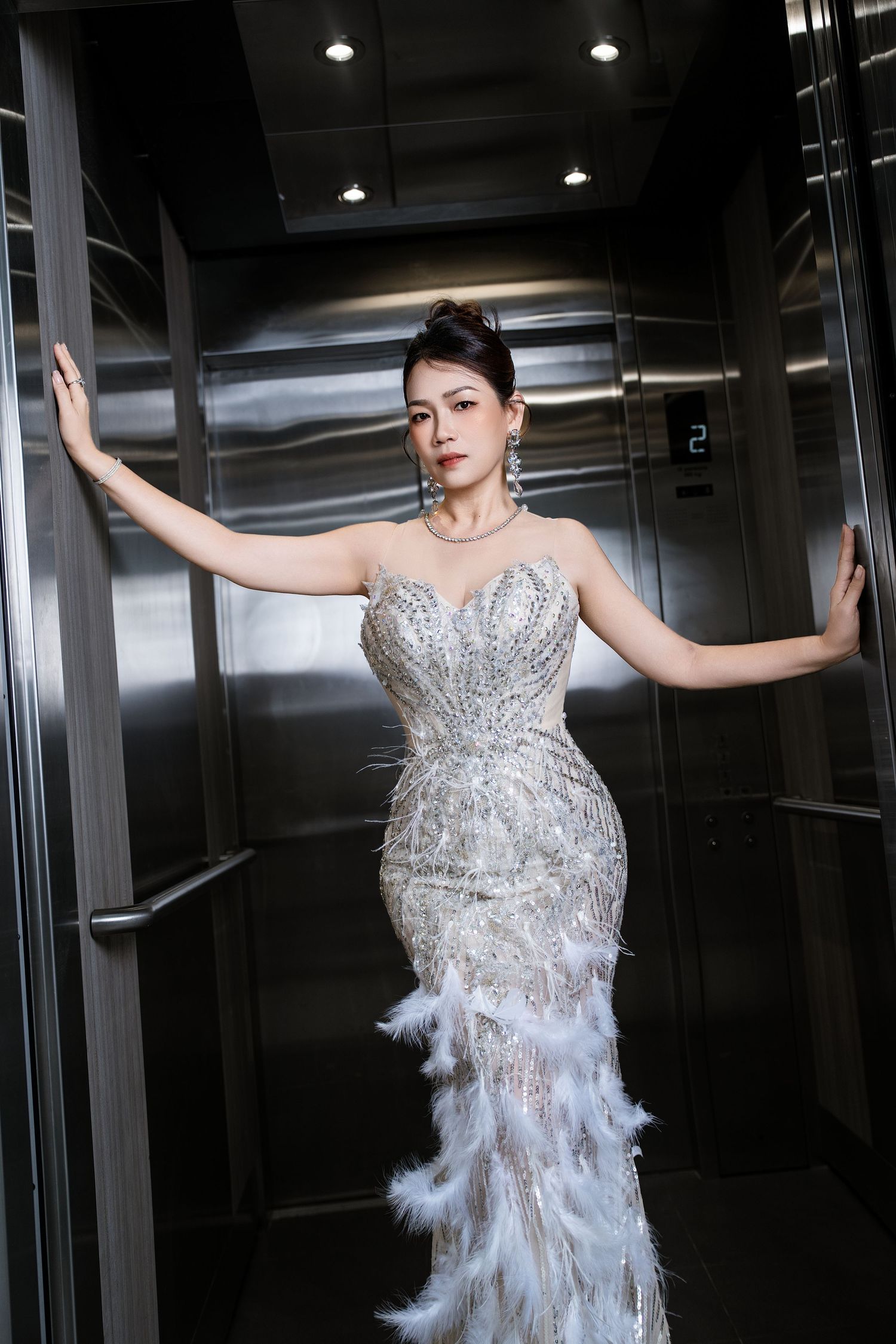 Woman in elegant beaded mermaid gown with feather hem posing in a silver elevator.