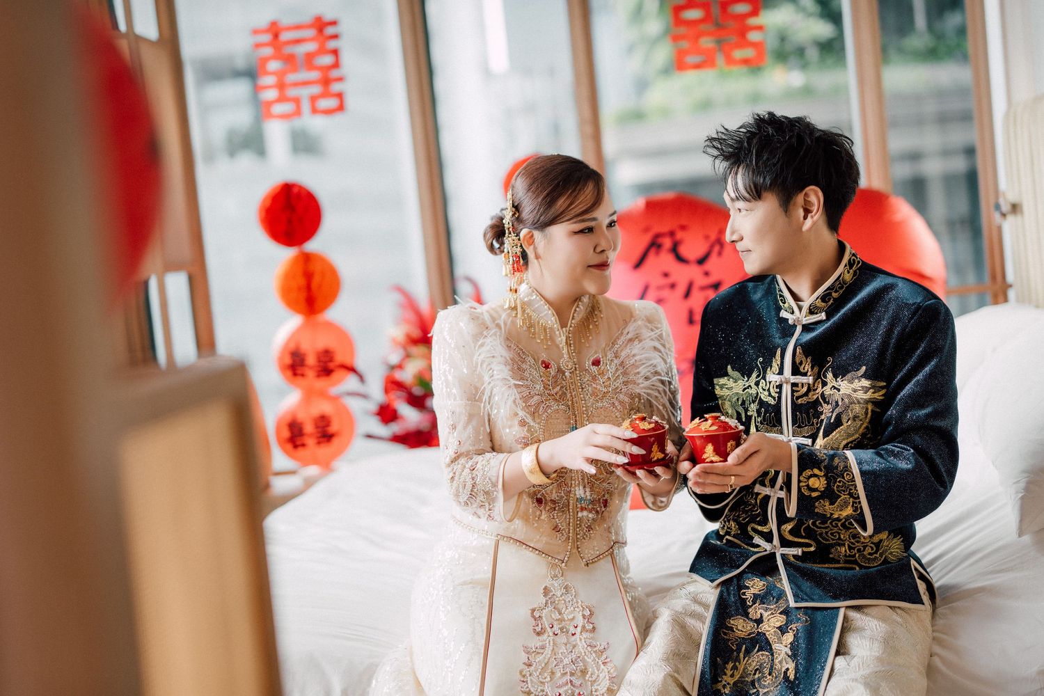 Couple in traditional Chinese wedding attire holding red gift boxes, with festive decorations in the background.