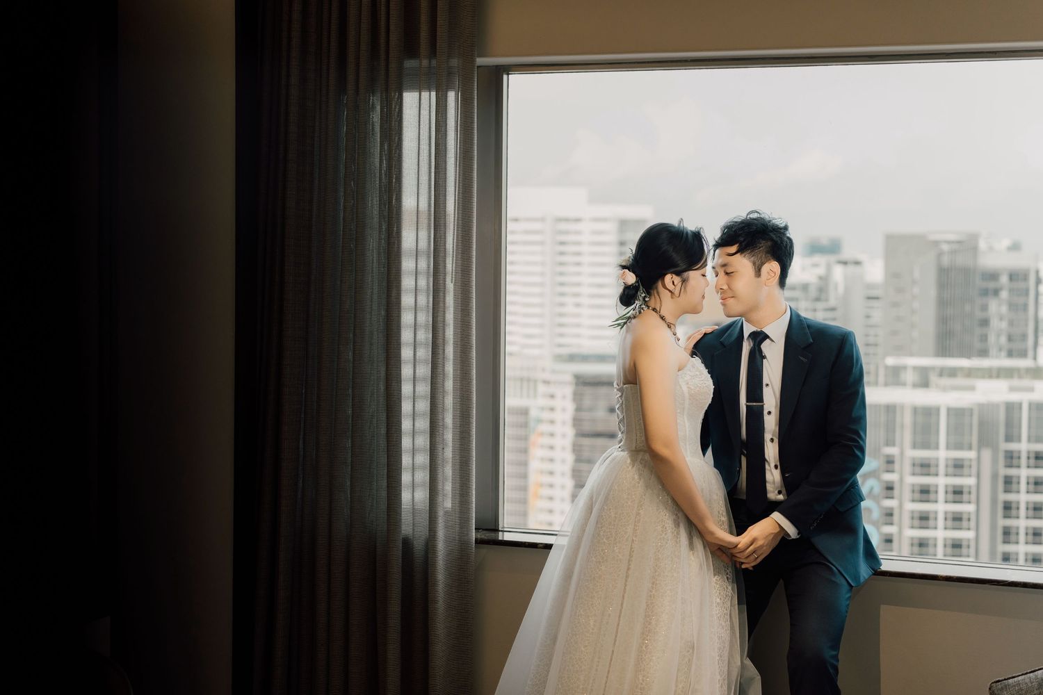 A couple shares a romantic moment near large windows with a cityscape view in the background.