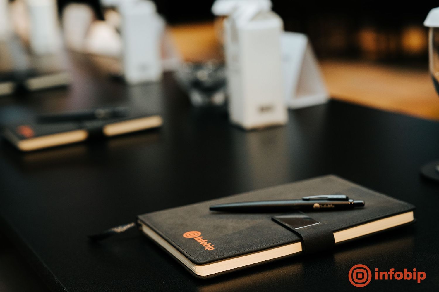 Black Infobip branded notebook with pen on dark conference table, with white name cards blurred in background.