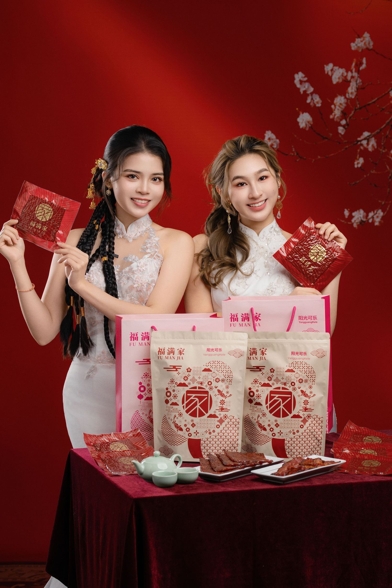 Two women in white cheongsam holding red envelopes beside Chinese New Year gift bags on a festive red background.