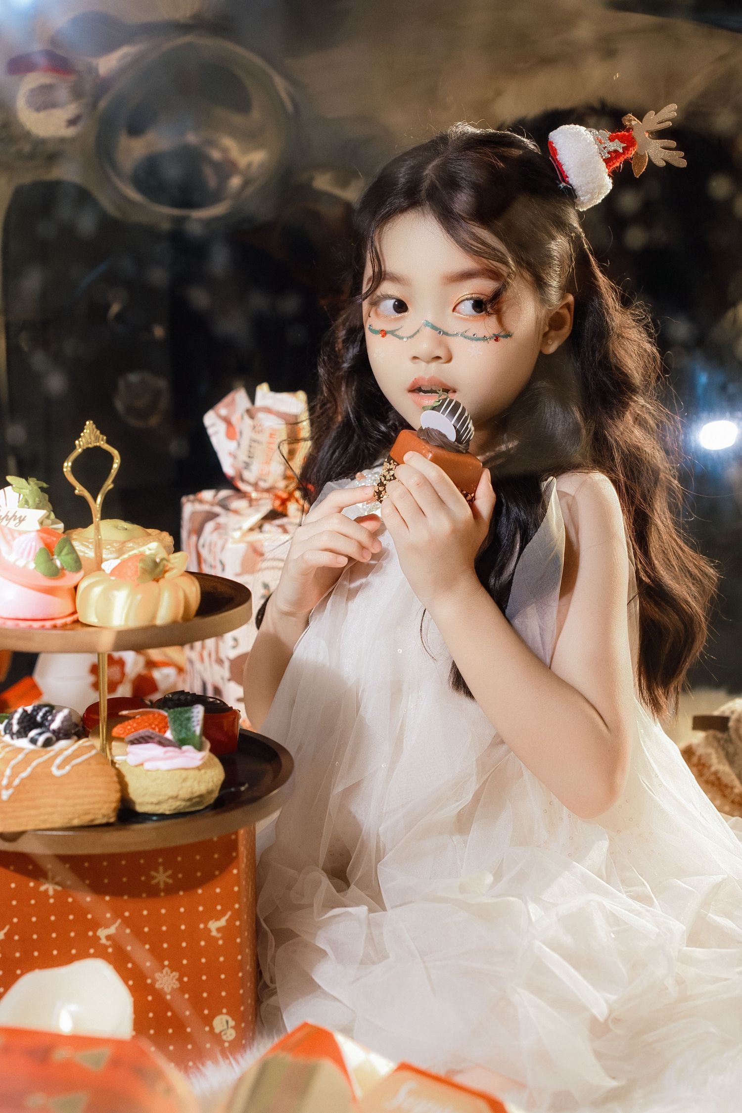 Young girl in white dress with face paint enjoys pastries at a Halloween-themed dessert display.
