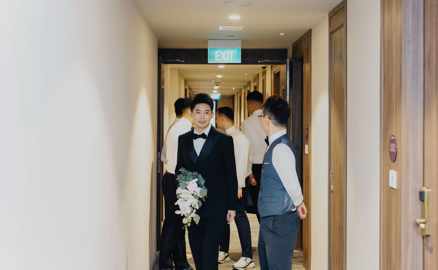 A narrow interior corridor with wooden doors and people gathered in formal attire for a special occasion.
