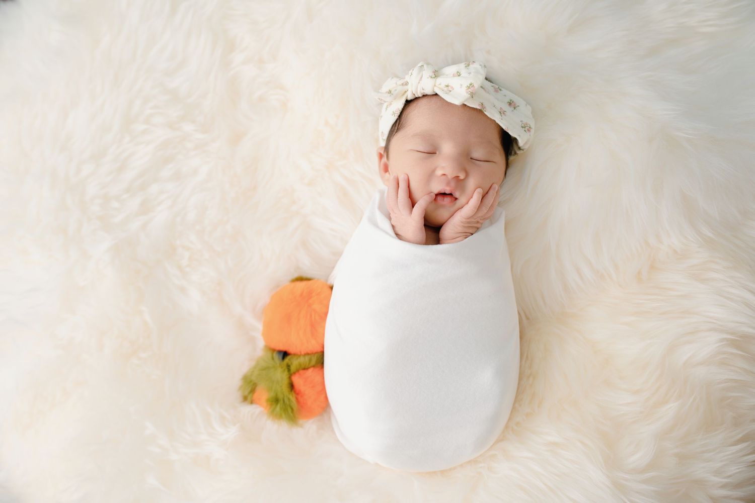 Newborn baby girl swaddled in white wrap with floral headband sleeping on fluffy white fur with mini pumpkin.
