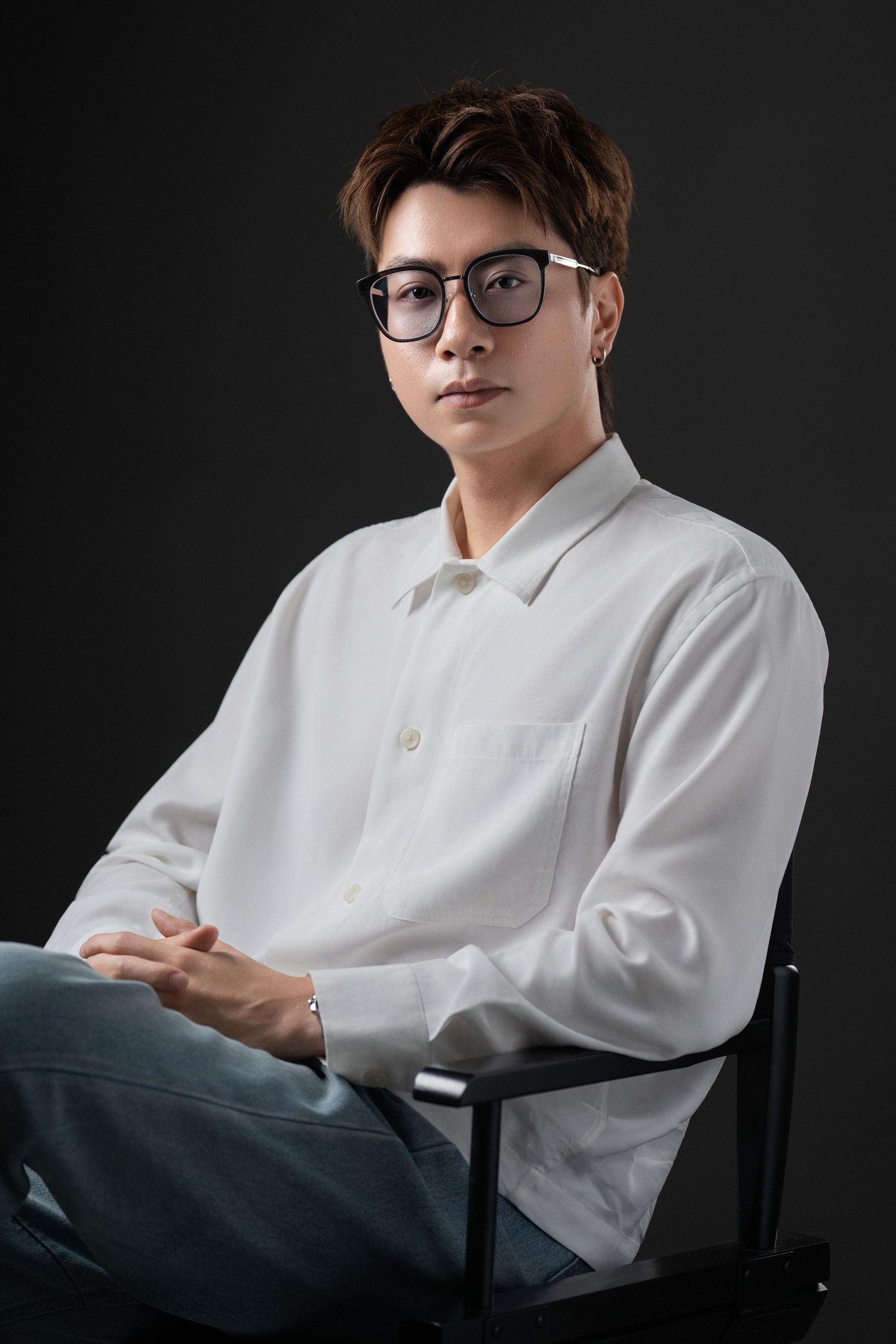Young Asian man in white shirt and glasses sitting in chair against dark background.