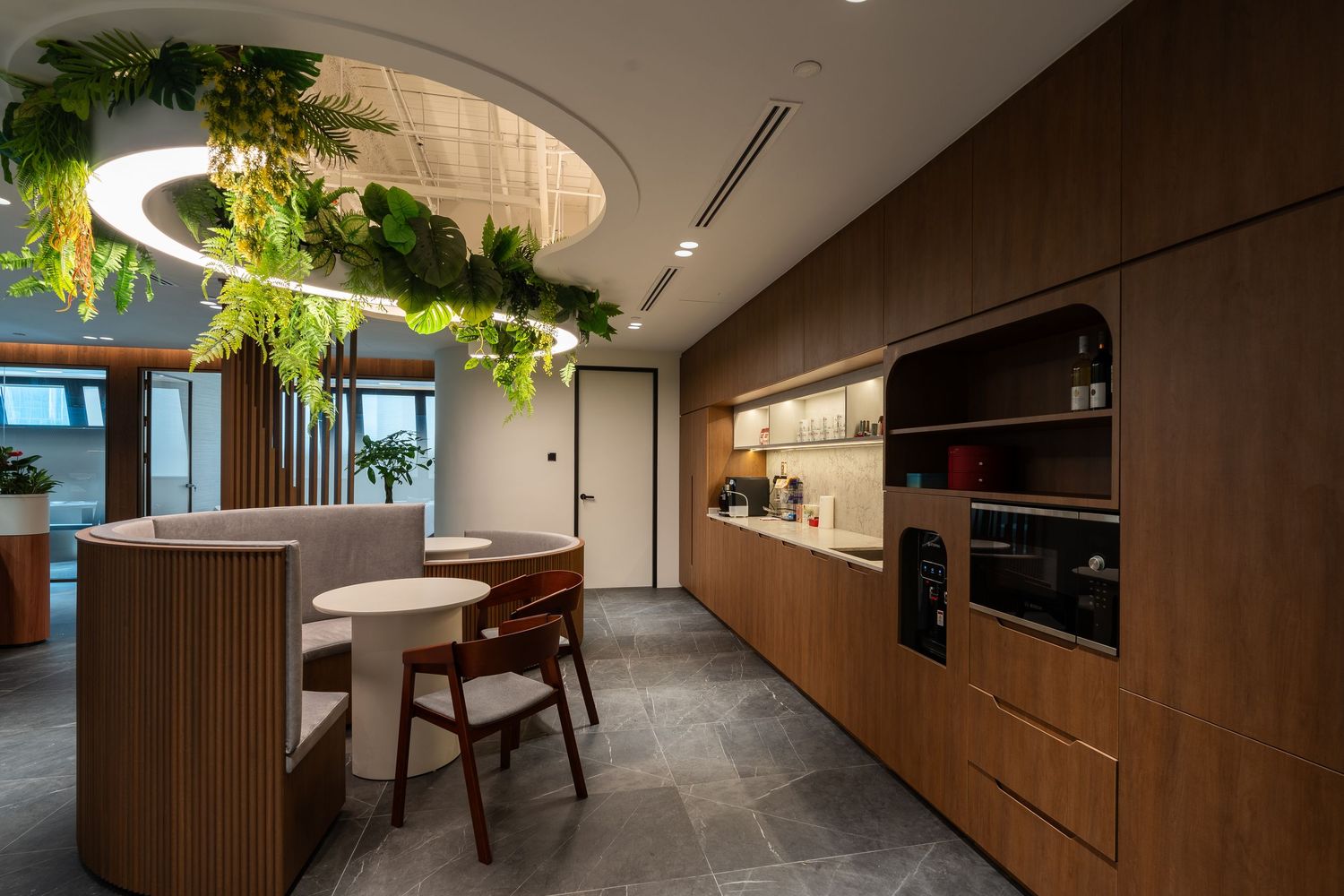 Modern office kitchen with wooden cabinetry, circular dining nook, and lush hanging plants under a round ceiling light.