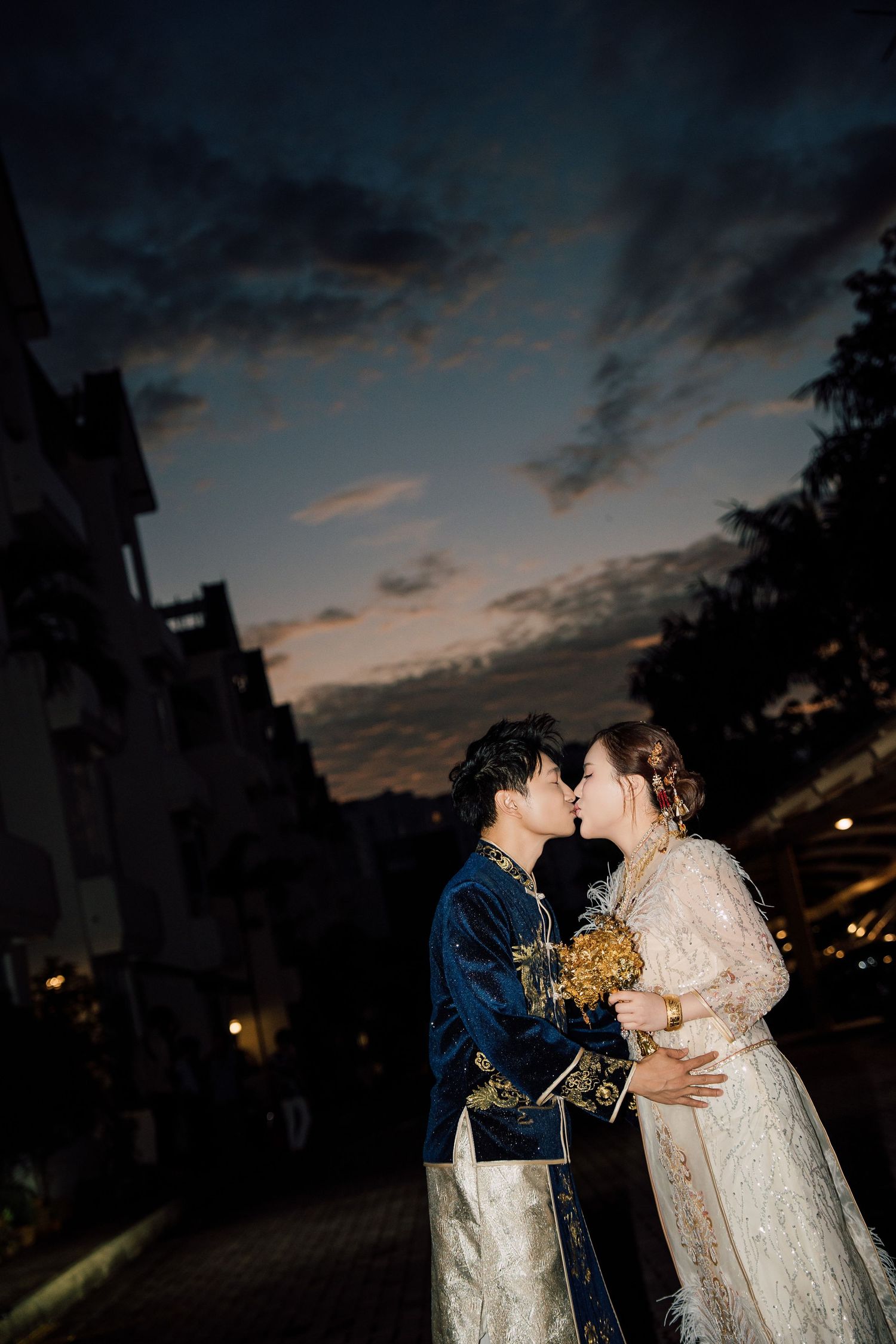 A couple in wedding attire shares intimate moments at sunset against a moody cloudy sky backdrop.