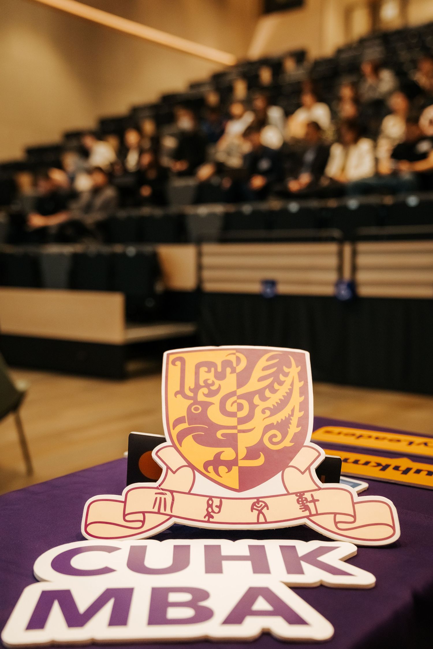 CUHK MBA logo and crest displayed on a purple table in a lecture hall with students seated in the background.