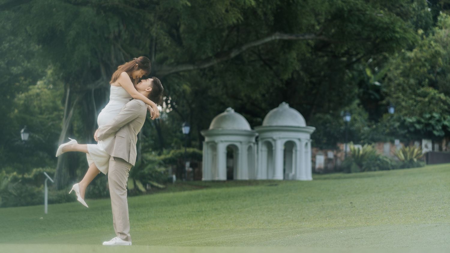 Couple sharing romantic embrace on lush green lawn with white gazebo in background during outdoor wedding photoshoot.