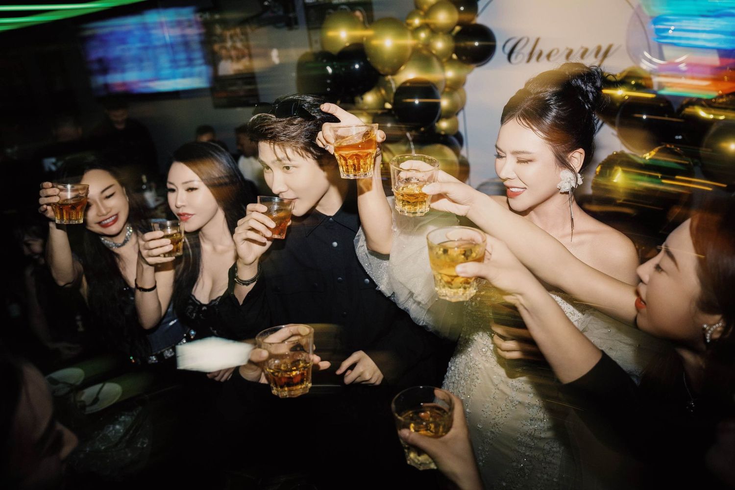 Group of young adults toasting with beer glasses at a lively nightclub party near a Cherry sign.