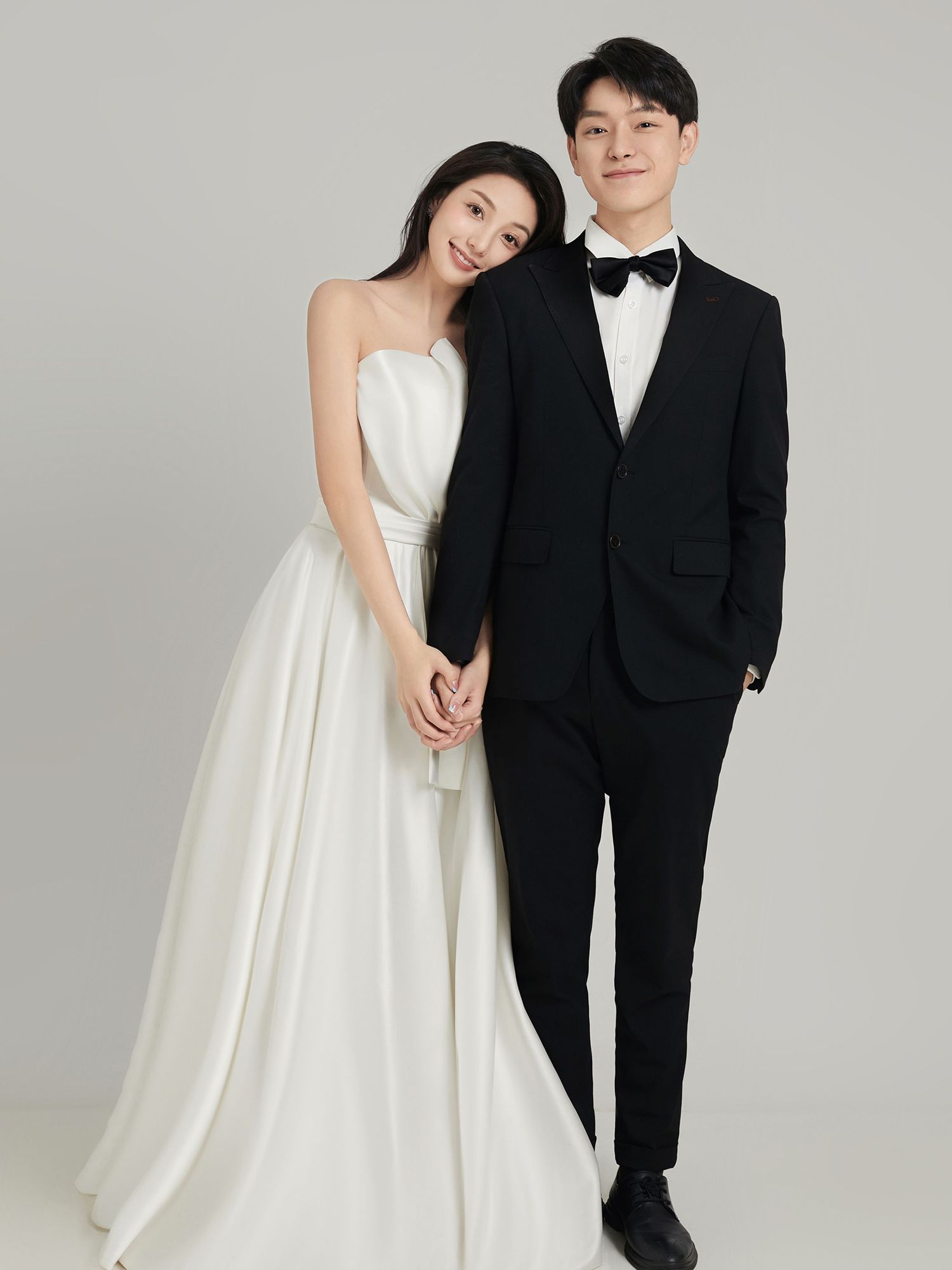 Couple posing for wedding portrait, woman in white strapless gown and man in black tuxedo with bow tie, holding hands.