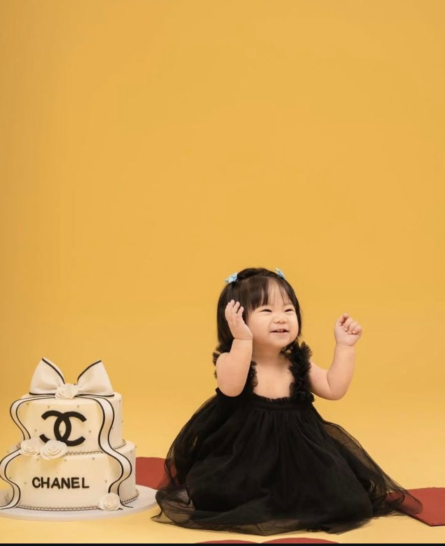 A toddler girl in a black dress sits beside a Chanel-themed cake against a yellow background.