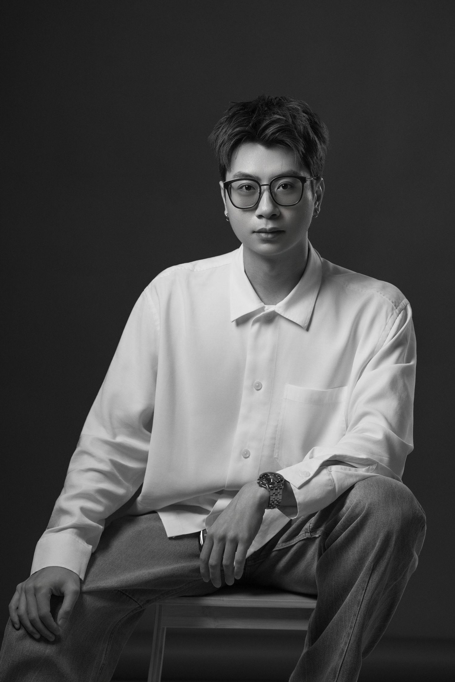 Black and white portrait of a young Asian man in white shirt and glasses, seated confidently in a studio setting.