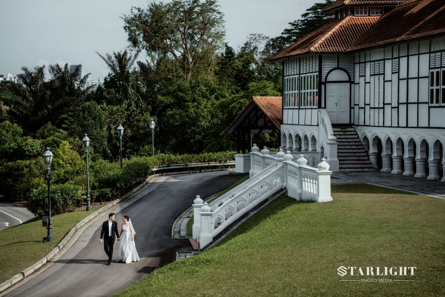 Couple in wedding attire walking along a scenic path beside a colonial white building surrounded by lush greenery.
