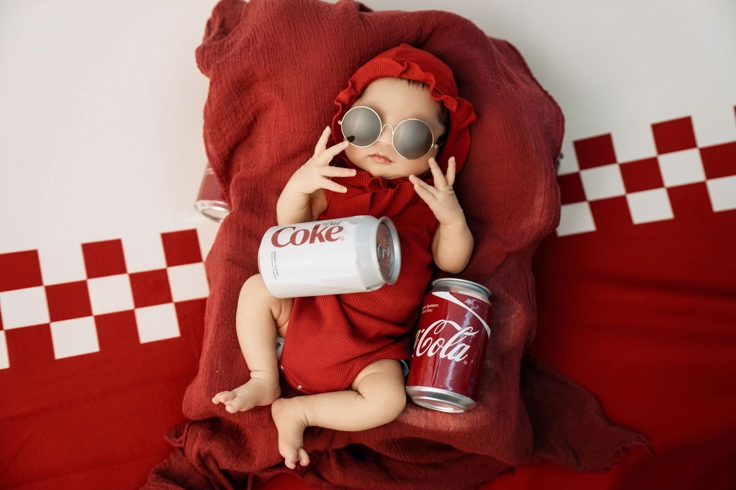 Baby in red outfit and sunglasses posed with Coca-Cola cans on red fabric backdrop.
