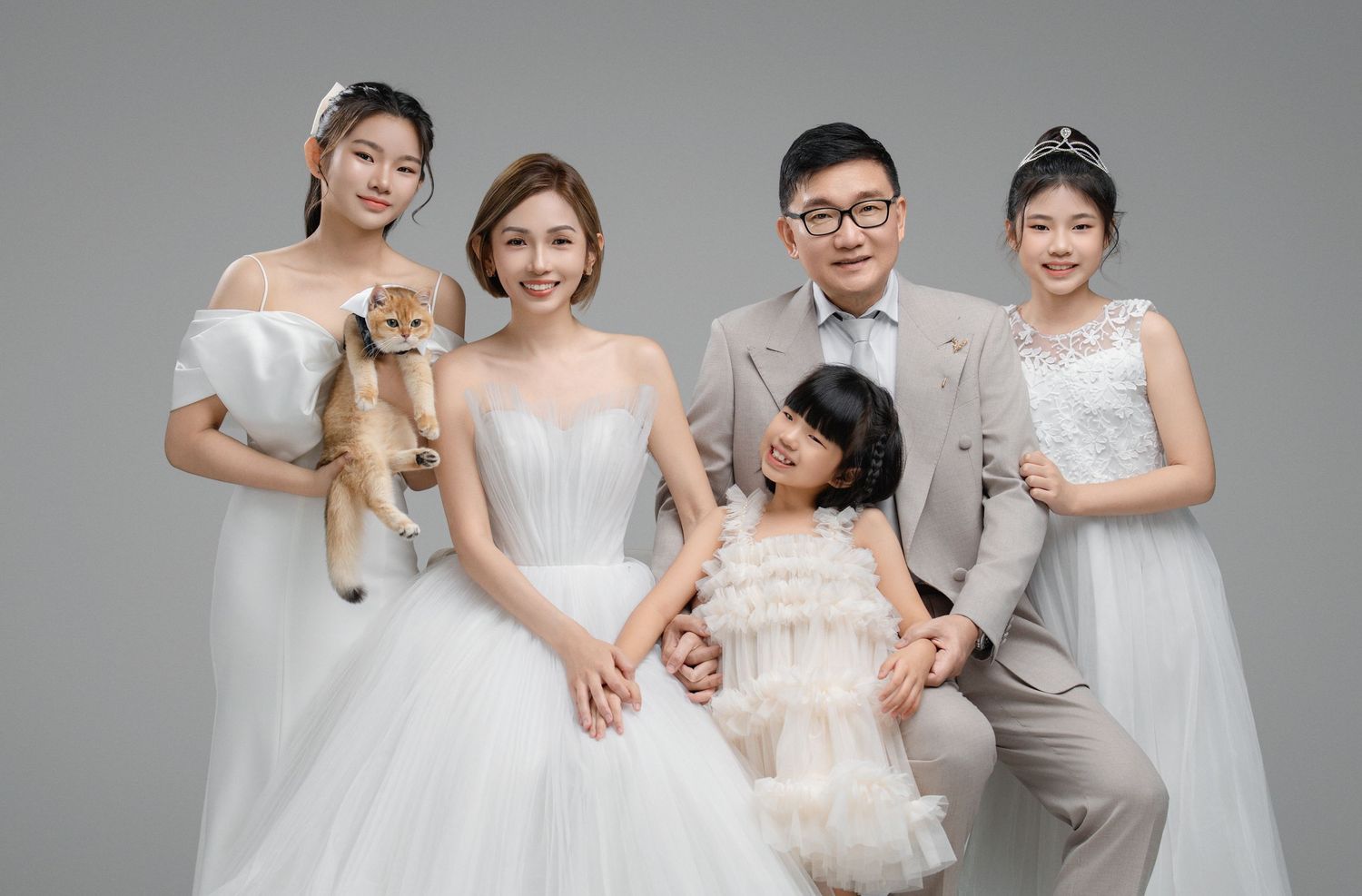 Family portrait with five people in elegant white and beige formal wear, holding a cat, posed against a gray studio background.