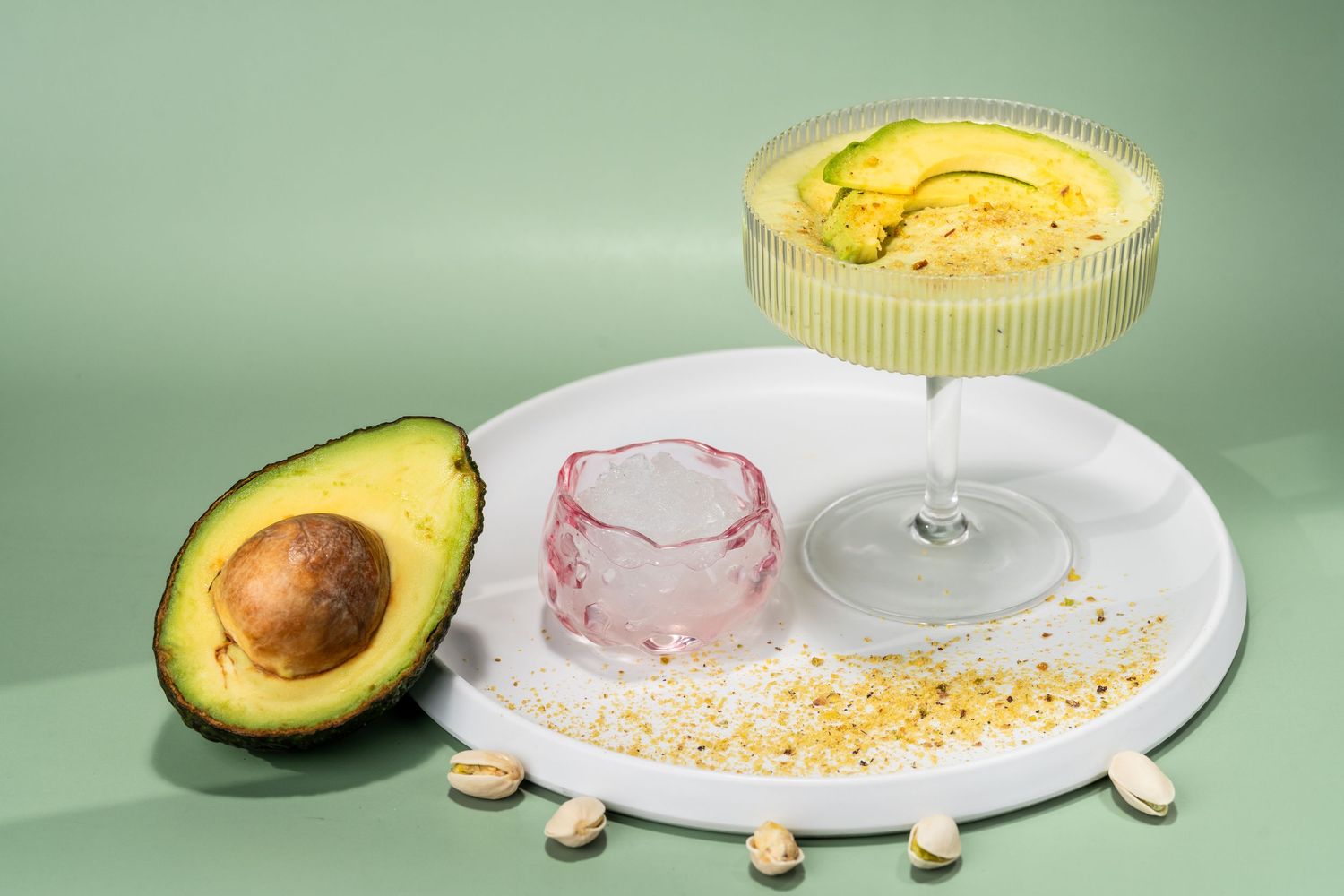 Avocado cocktail in coupe glass with pistachios and pink ice cube on white plate, green background.