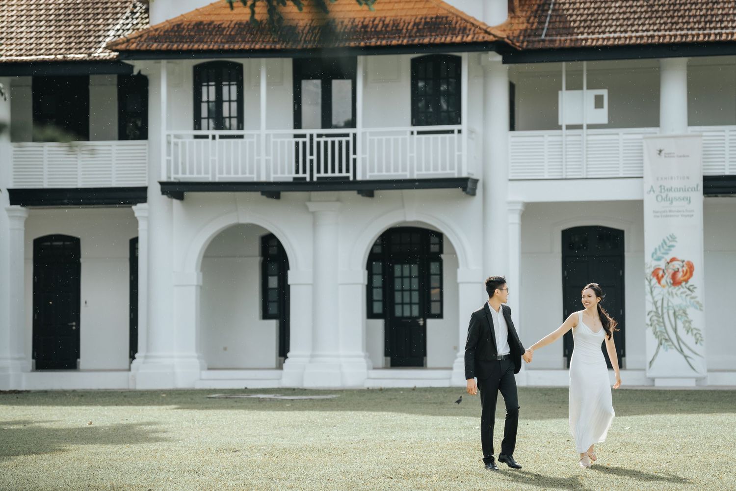 Couple in wedding attire holding hands walking in front of a white colonial building with arched windows.