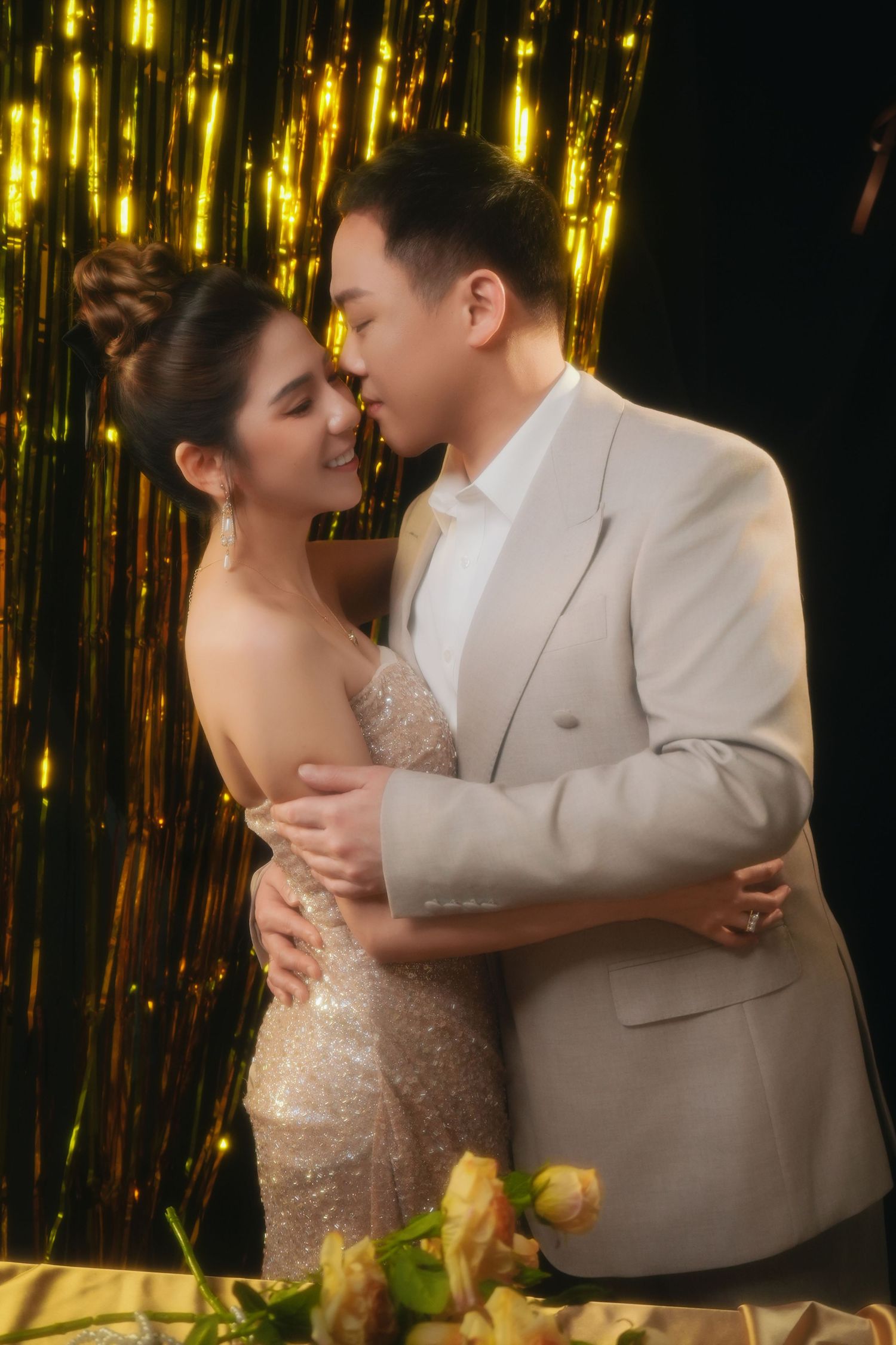 Romantic couple embraces in front of shimmering gold backdrop during formal celebration.