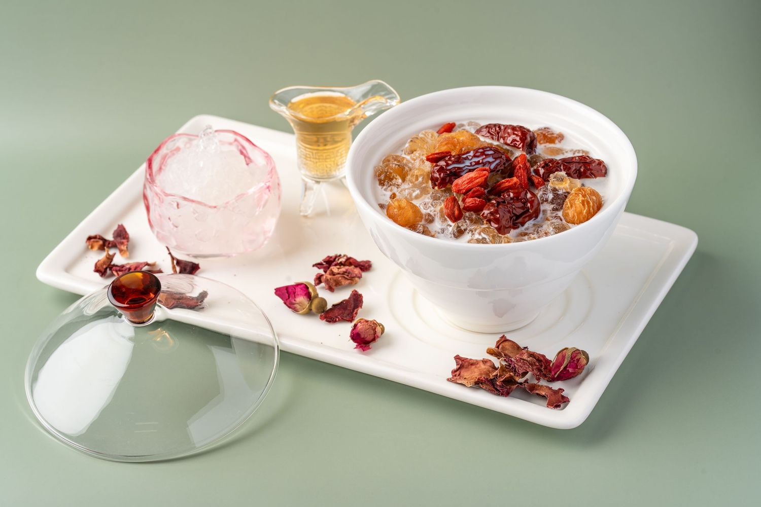 A white bowl of oatmeal topped with dried fruits and nuts on a tray with rose petals and a glass dome.