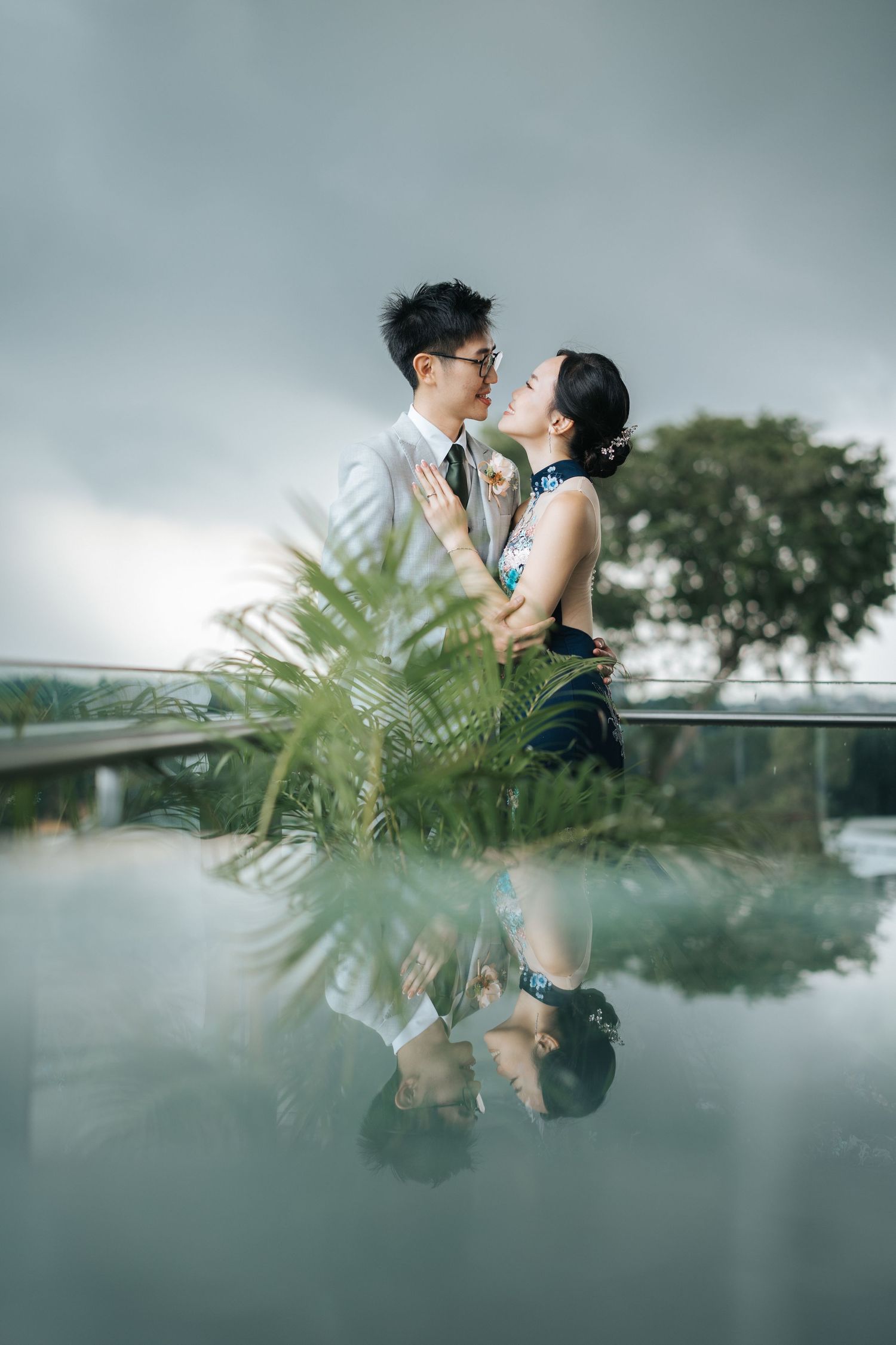 Couple in wedding attire sharing a romantic moment by a reflective pool with tropical plants and cloudy sky.