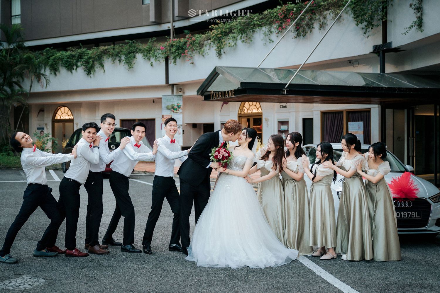 A wedding party strikes a fun pose outdoors, with bridesmaids in gold dresses and groomsmen in white shirts and dark pants.
