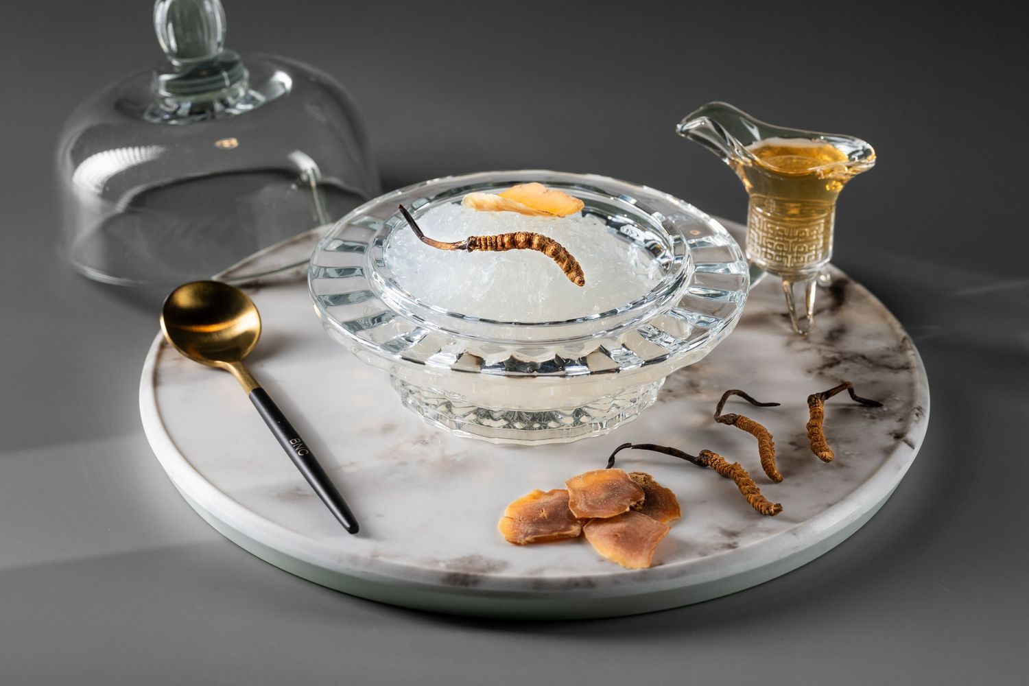 Elegant marble tray with crystal bowl, gold spoon, and glass cloche featuring dried fruits and gourmet garnishes.