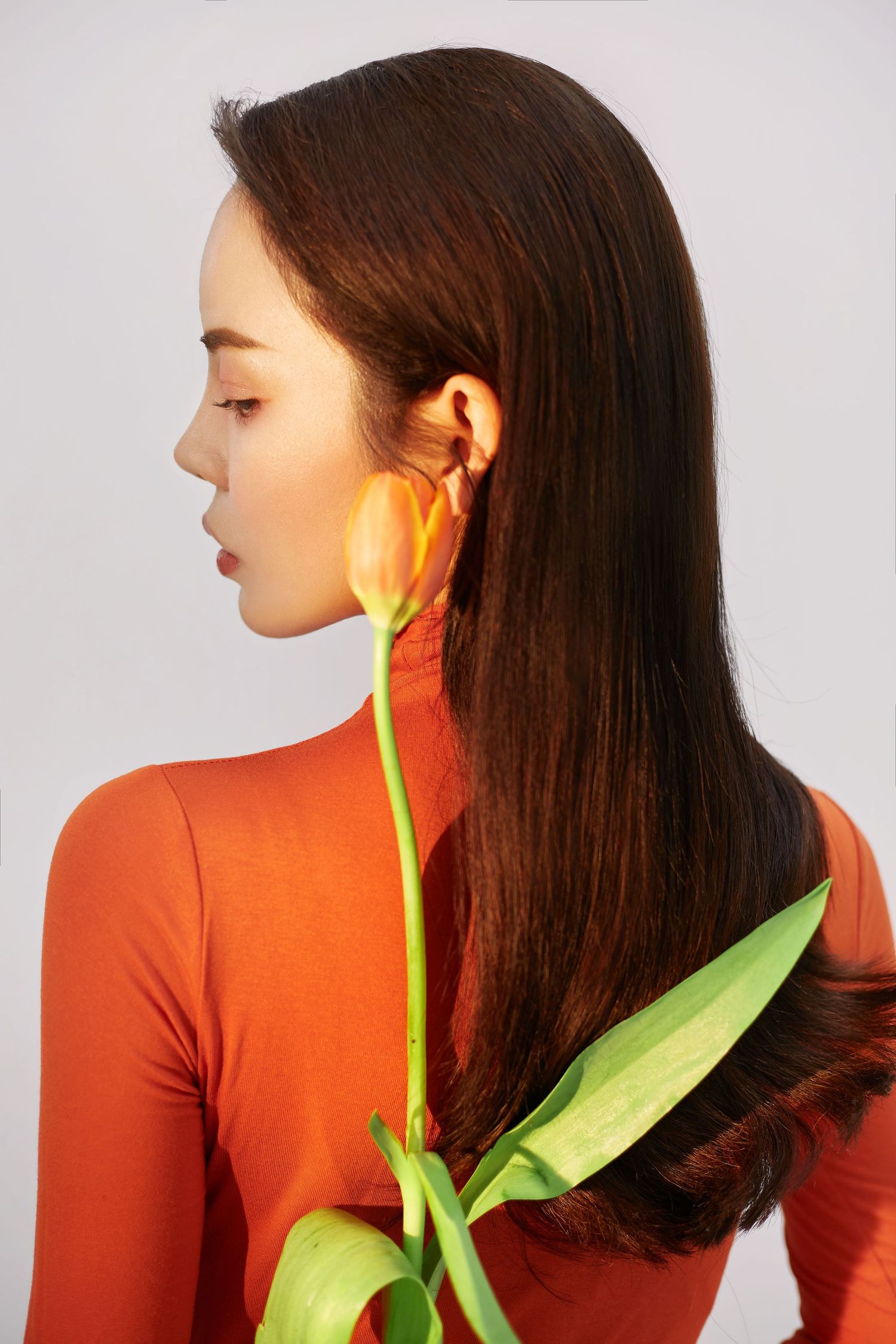Profile view of person with long dark hair wearing orange top posing with yellow tulip against white background.